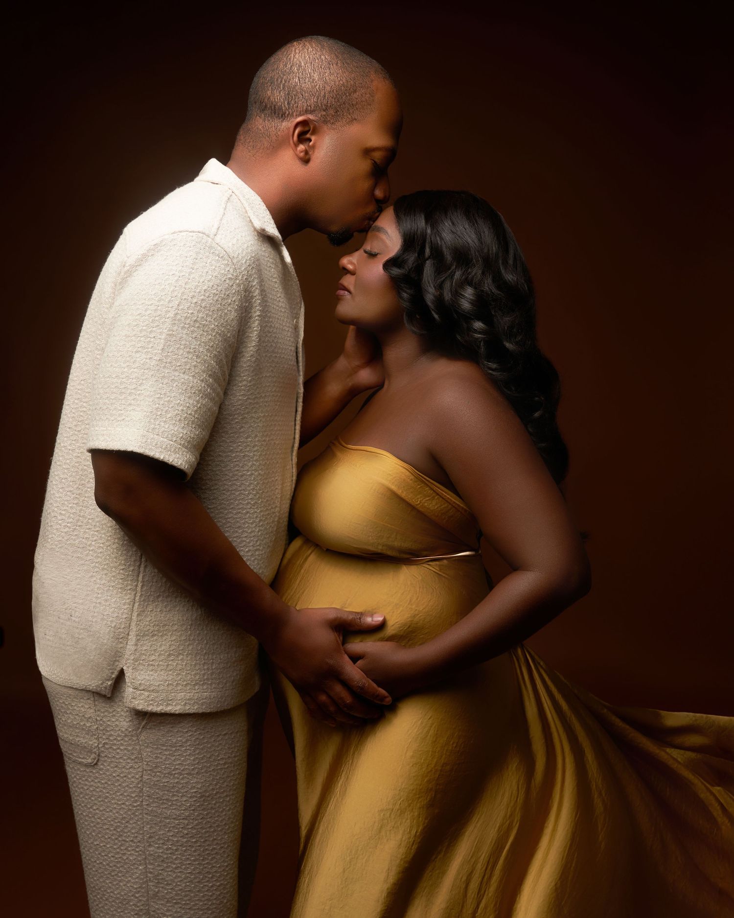 Romantic maternity photos with couple in gold dress and cream suit against dark background.