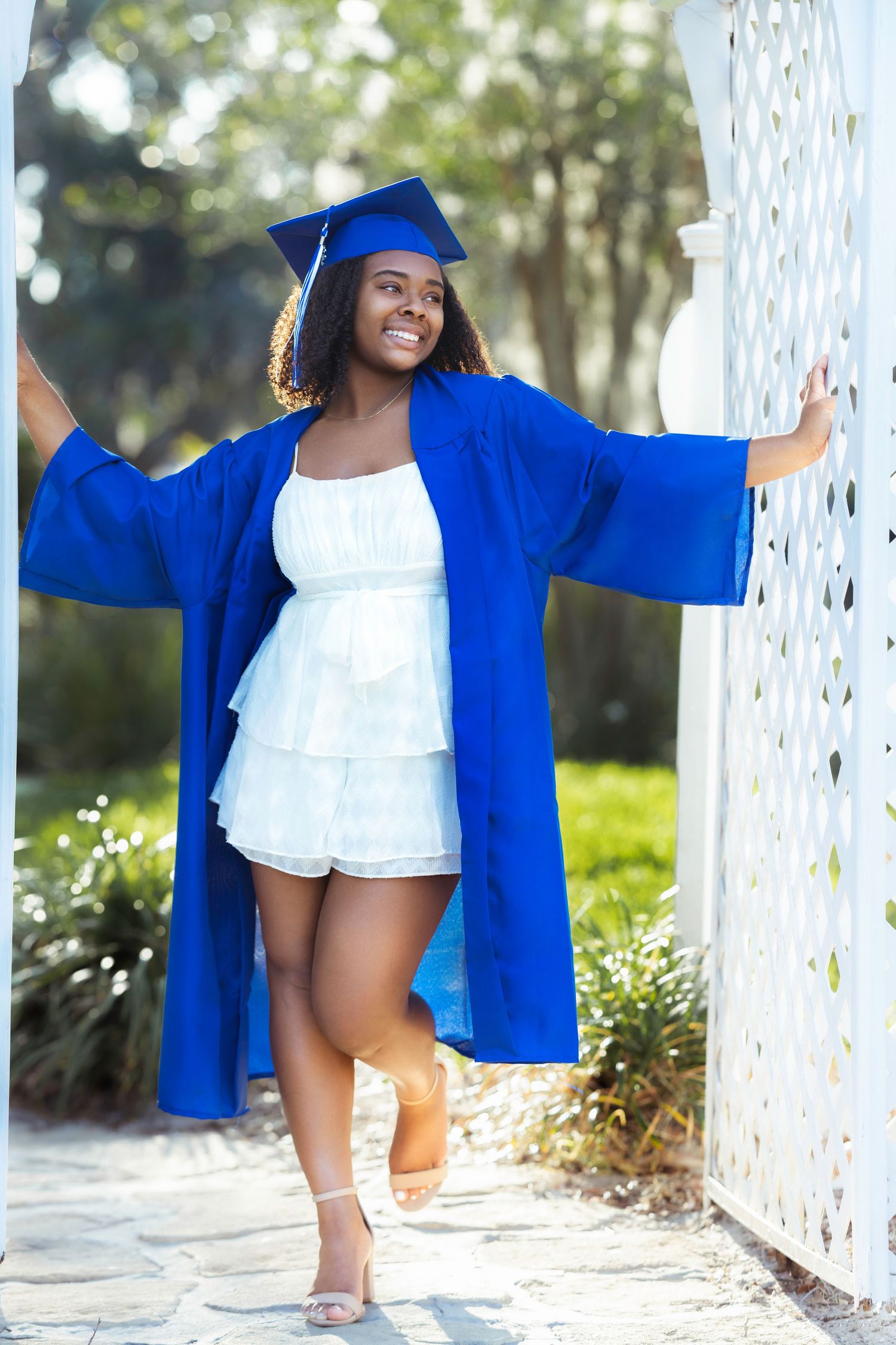 Graduate in royal blue cap and gown poses playfully between white pillars in an outdoor setting.