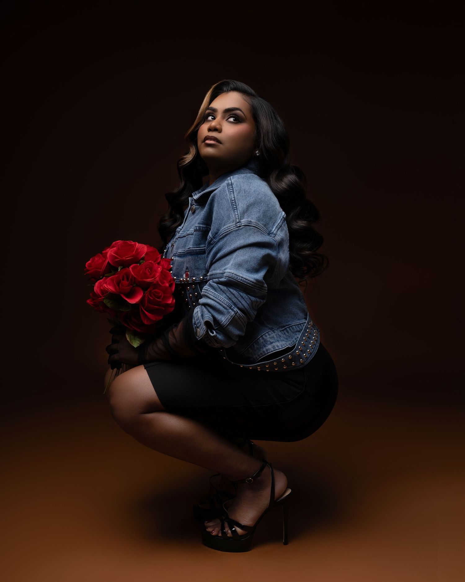 A person in denim jacket crouches in dramatic lighting while holding vibrant red roses against a dark background.
