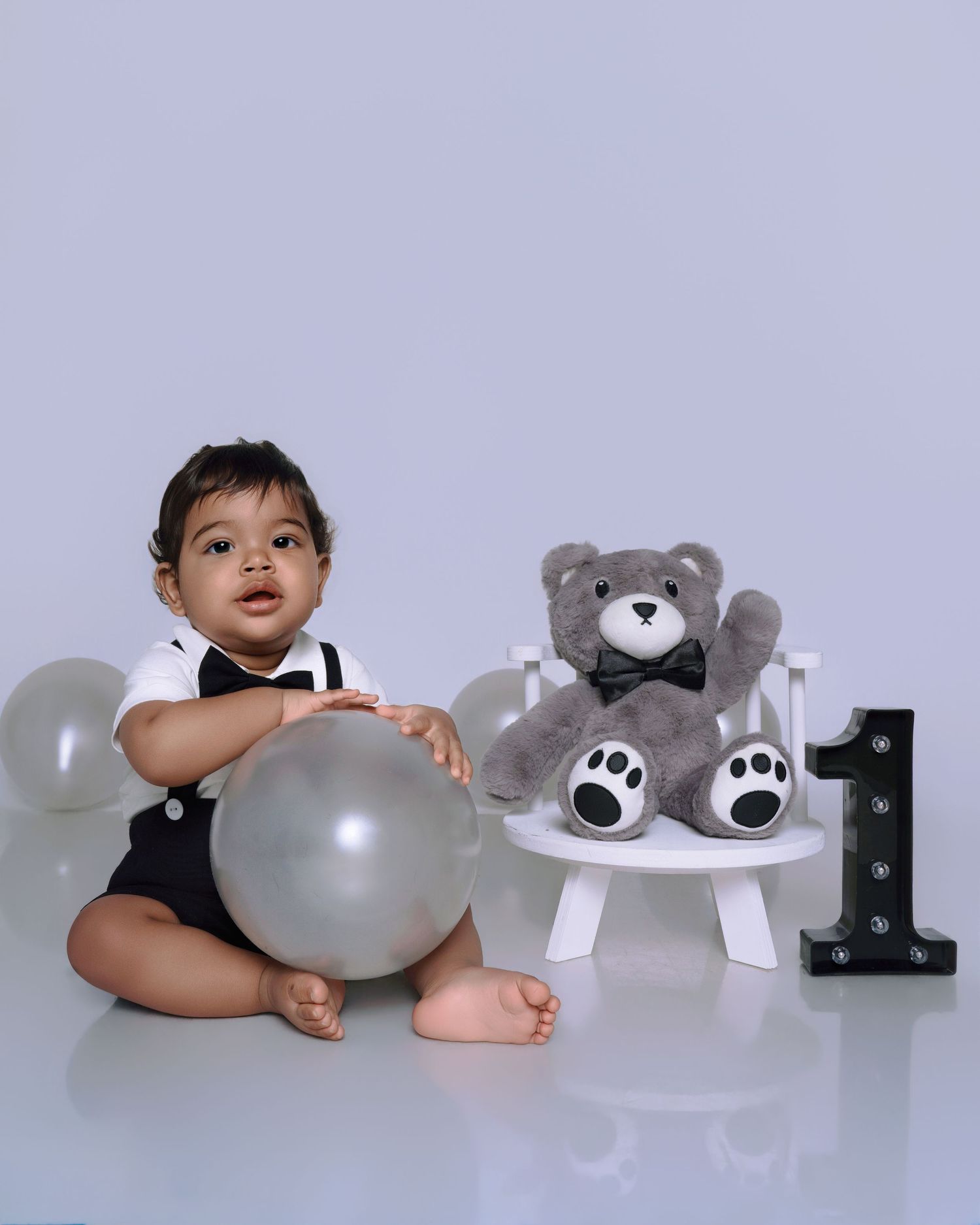 Baby boy in tuxedo romper sits with silver balloon beside teddy bear and number 1 sign for first birthday photo.