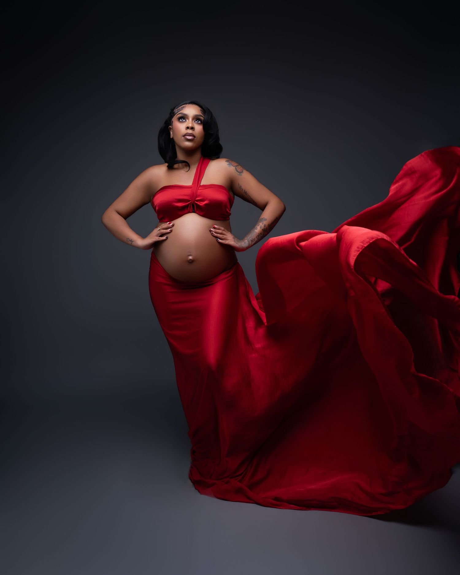 Pregnant woman in flowing red halter gown with dramatic fabric movement against dark studio background.