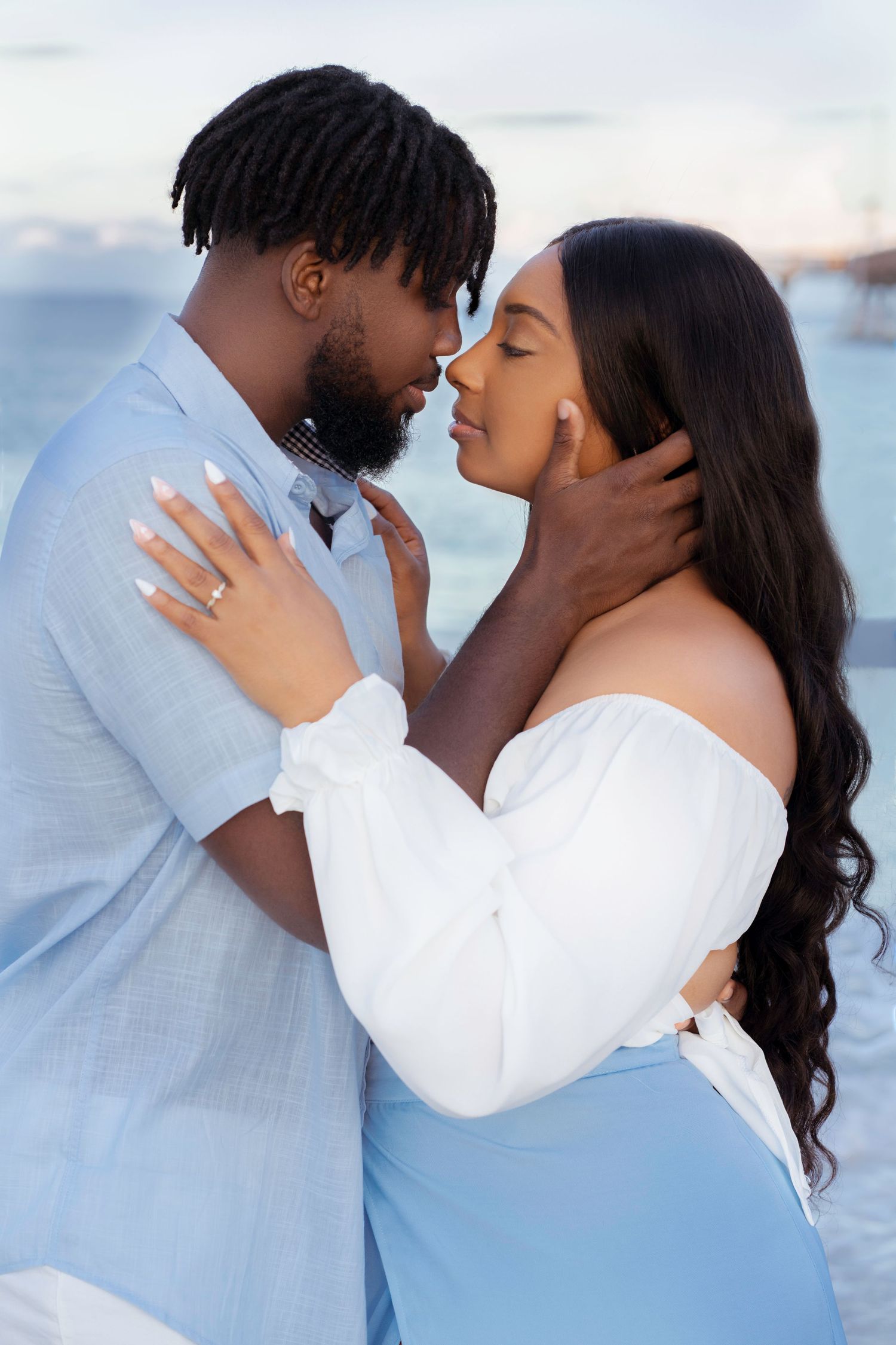 A romantic couple in white and light blue outfits share an intimate moment against a waterfront backdrop at sunset.