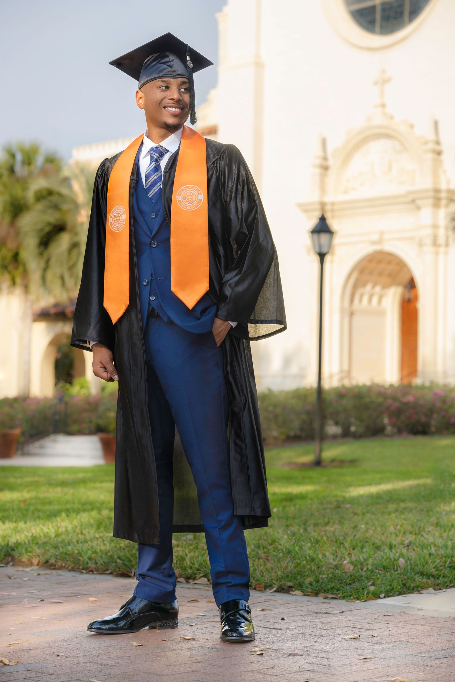 Graduate in cap and gown with orange stole photographed in front of a building.