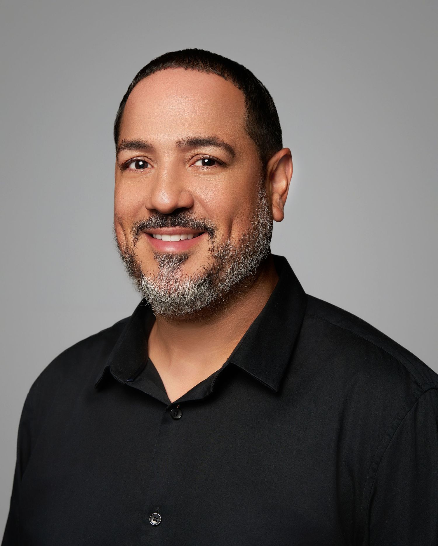 Professional headshot of someone wearing a black polo shirt against a gray background, with a warm smile and salt-and-pepper beard.