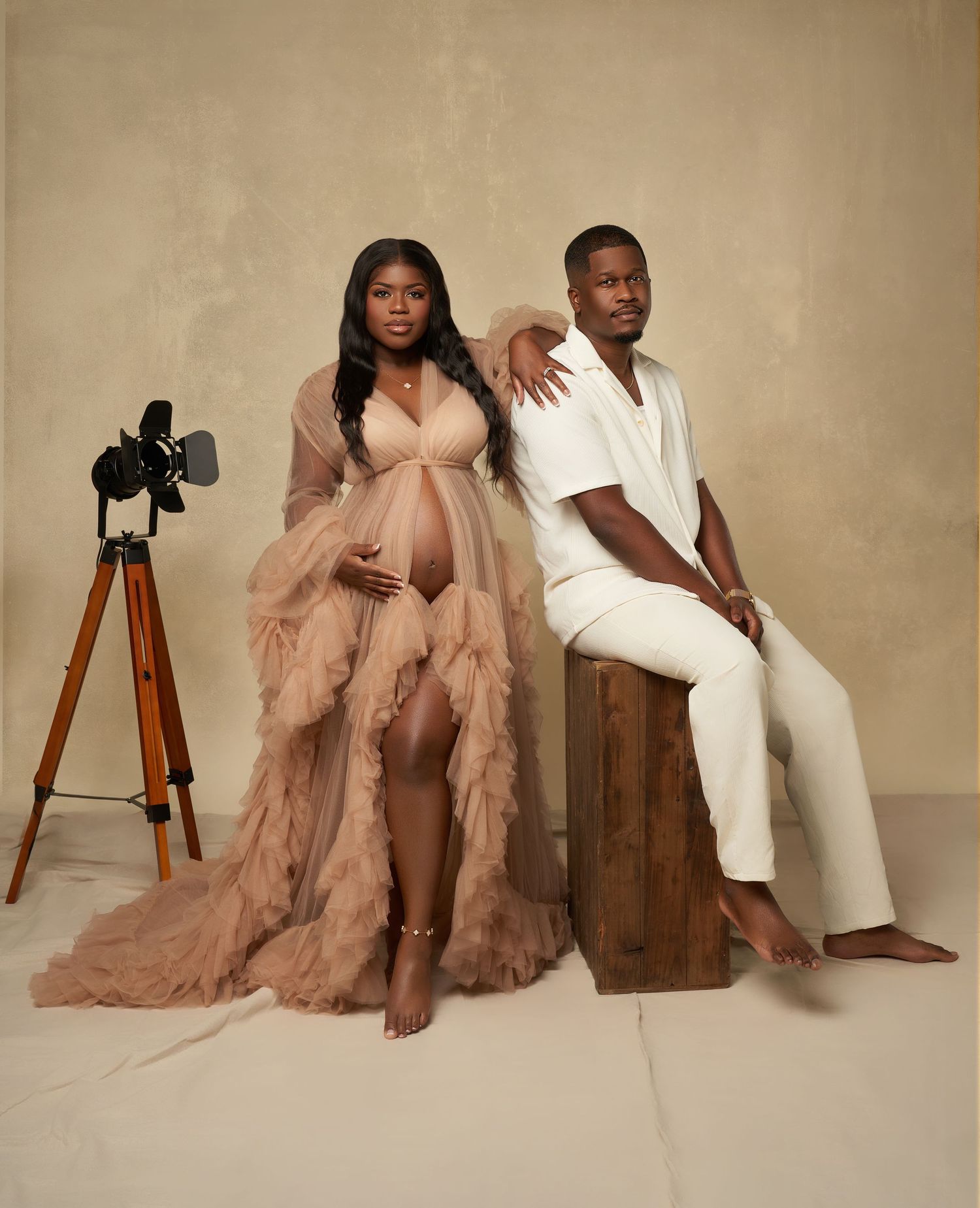 Stylish maternity session with couple posing on wooden furniture in neutral tones and flowing dress.