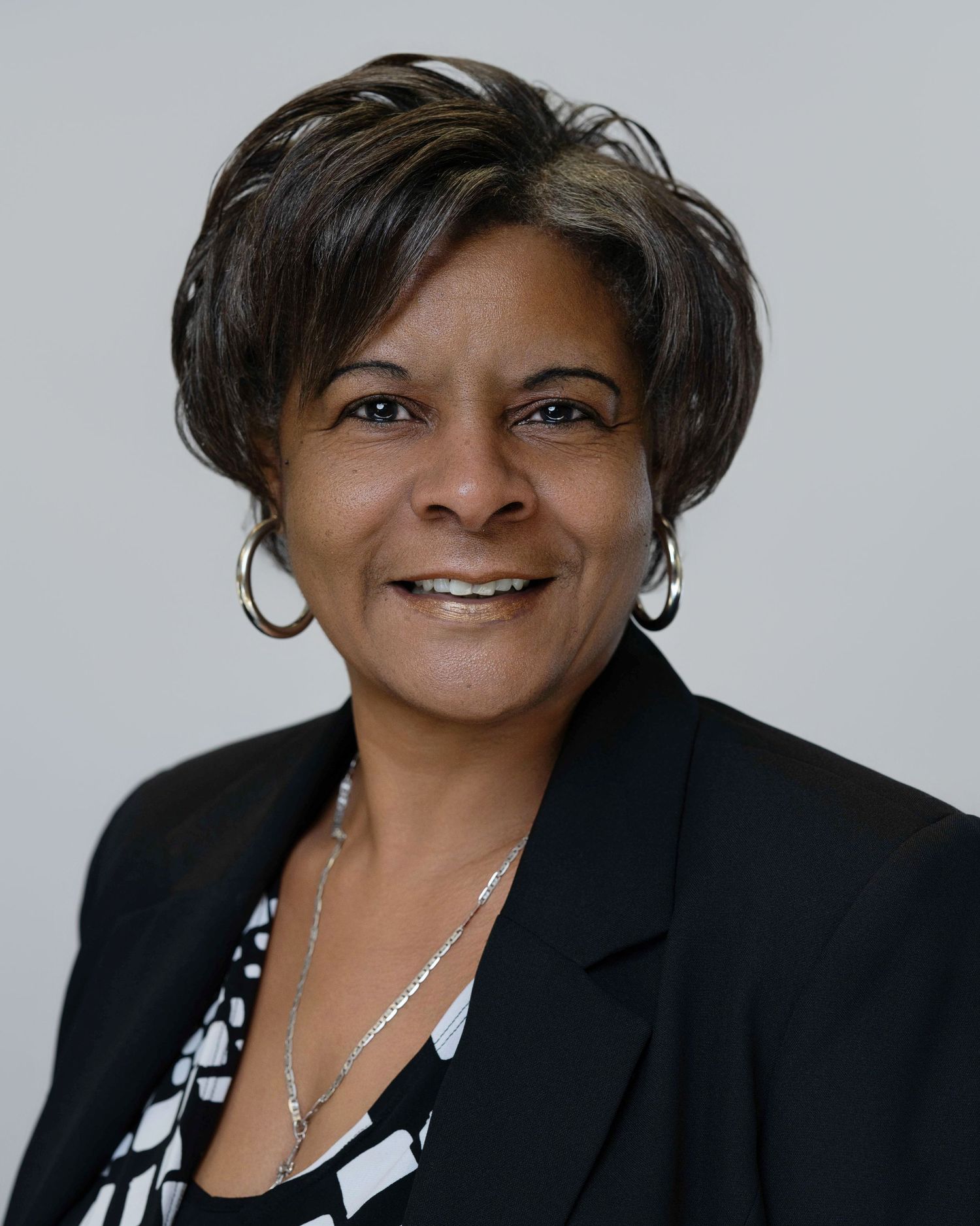 Professional headshot of someone in a black blazer and patterned blouse smiling warmly against a light background.