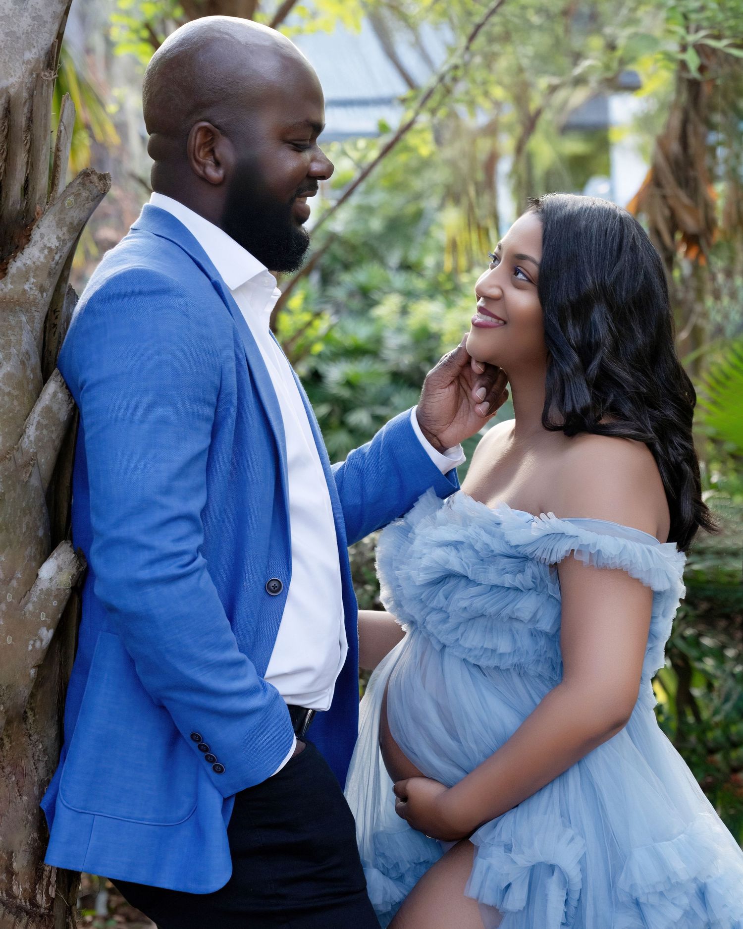 Couple in maternity photoshoot, man in blue suit tenderly touches pregnant woman's chin outdoors.