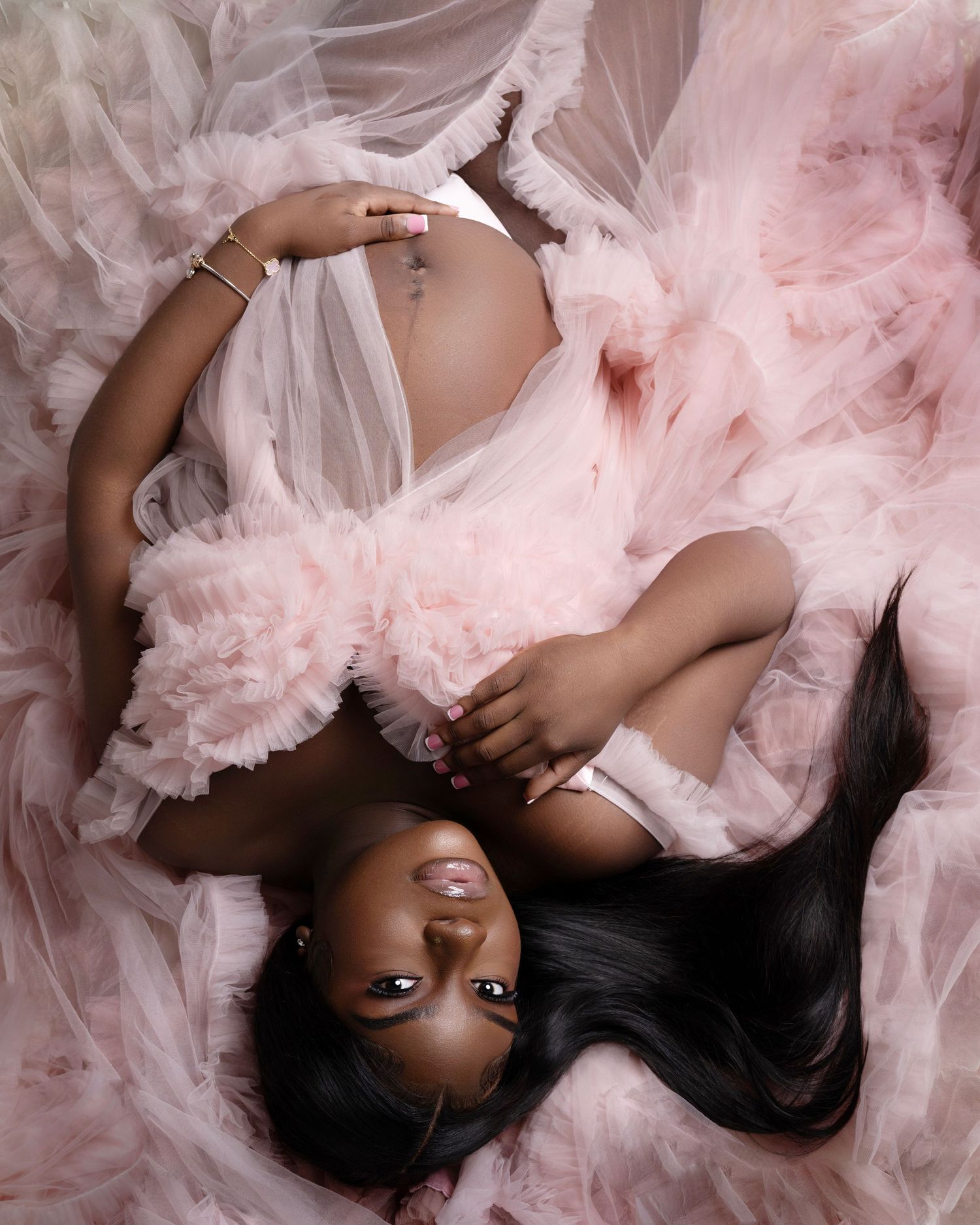 Pregnant woman in pink tulle robe lying down, showcasing baby bump in elegant maternity photography session.