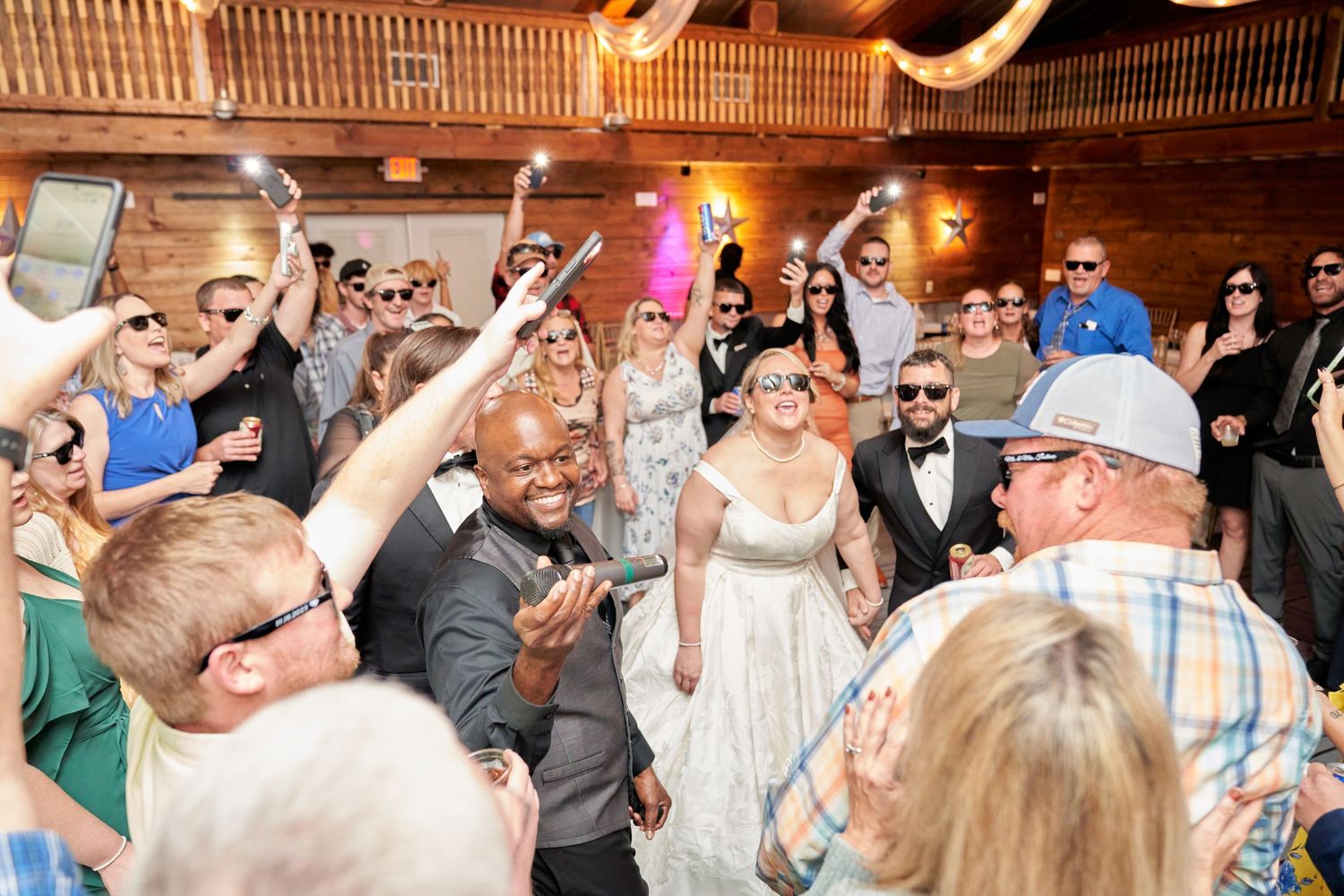 Wedding guests celebrate and dance together during a lively reception in a rustic wooden venue with raised arms and drinks.