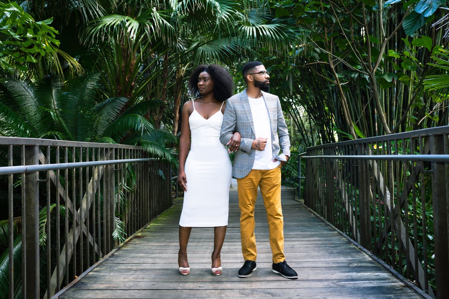Couple walking together on a wooden boardwalk path surrounded by lush tropical plants and palm trees.