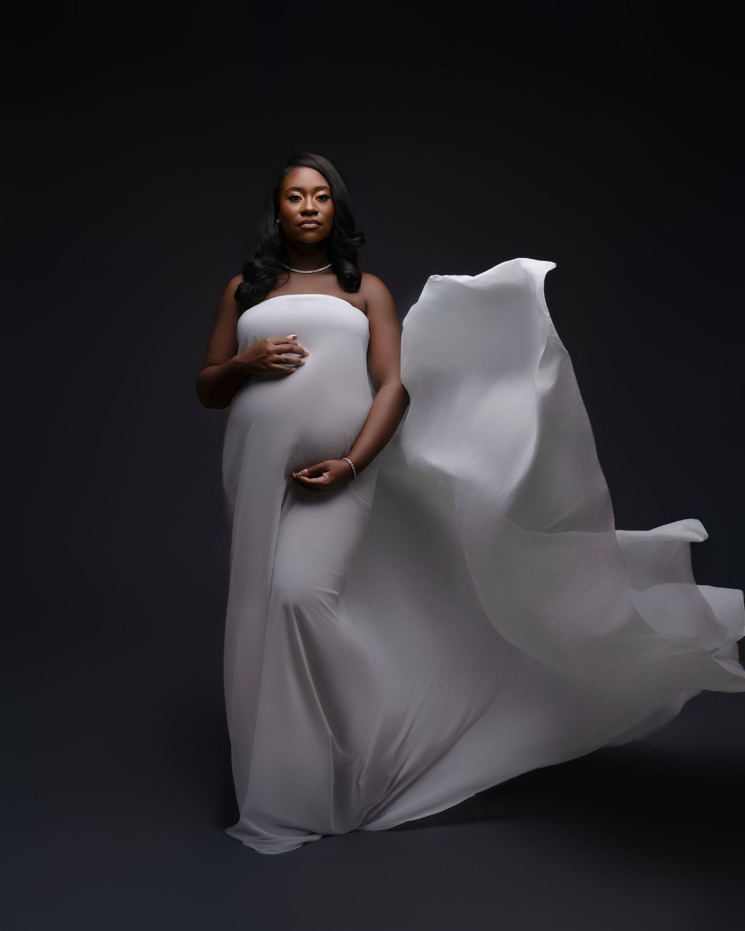 Artistic maternity photoshoot with flowing white fabric against dark background creating dramatic movement and contrast.