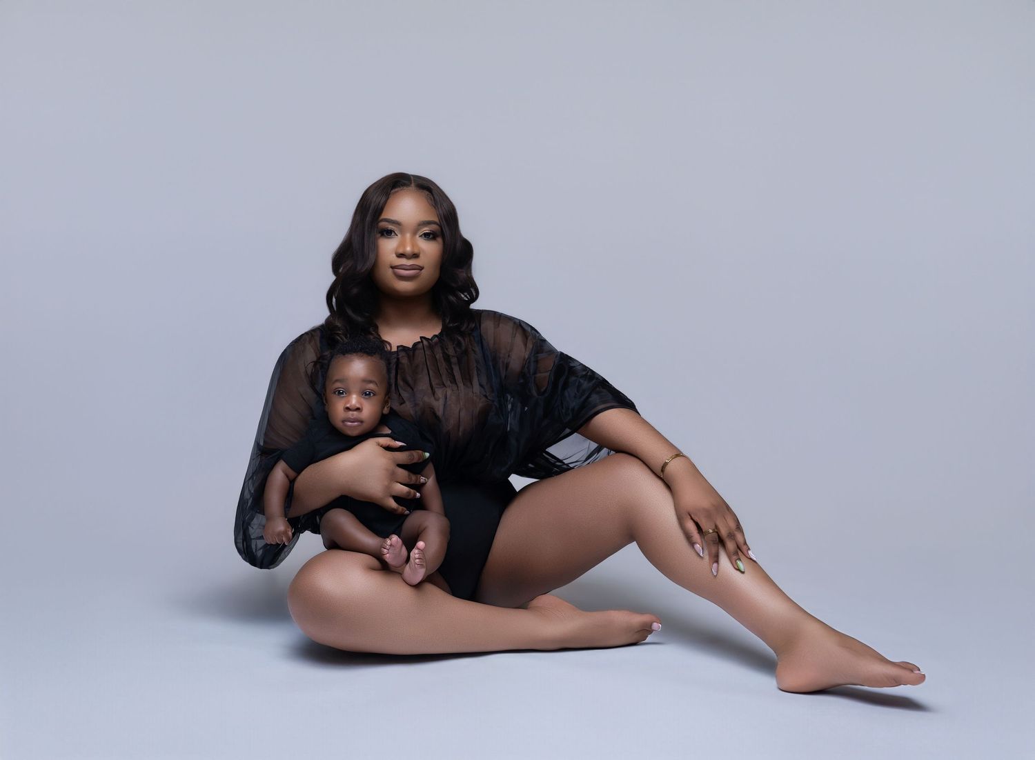 A woman in a black sheer bodysuit sits gracefully on a studio floor holding a baby in a professional portrait photo.