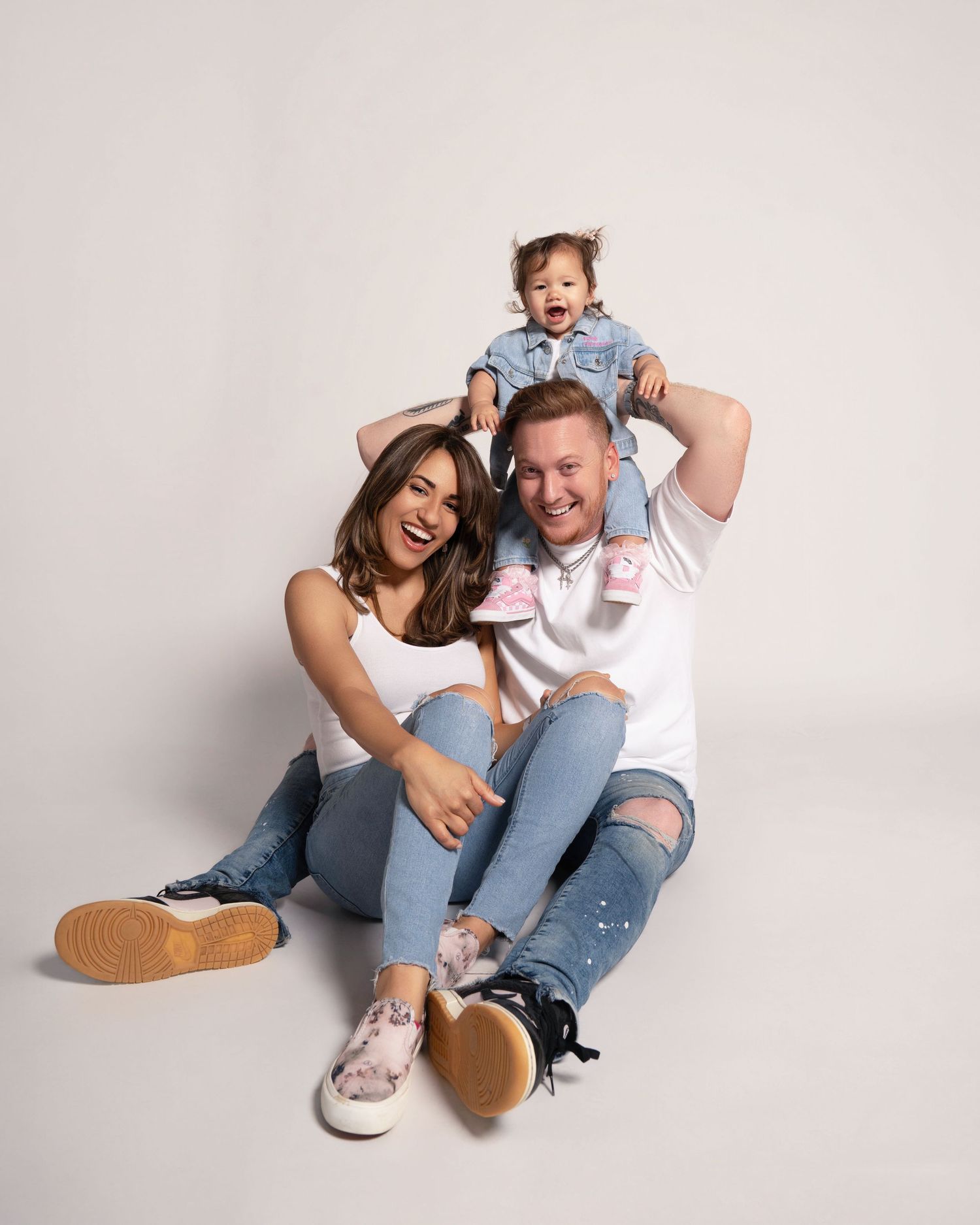 Family photo of a family sitting down and a baby sitting on the fathers shoulders against a white studio background
