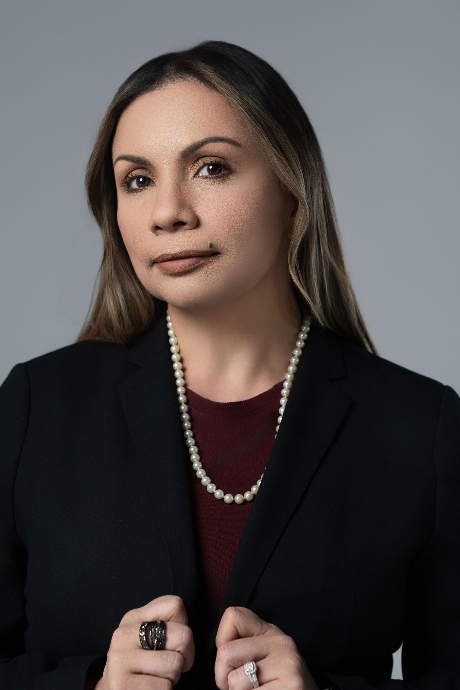 Professional studio portrait in dark tones featuring burgundy top, black blazer, and pearl necklace against gray background.