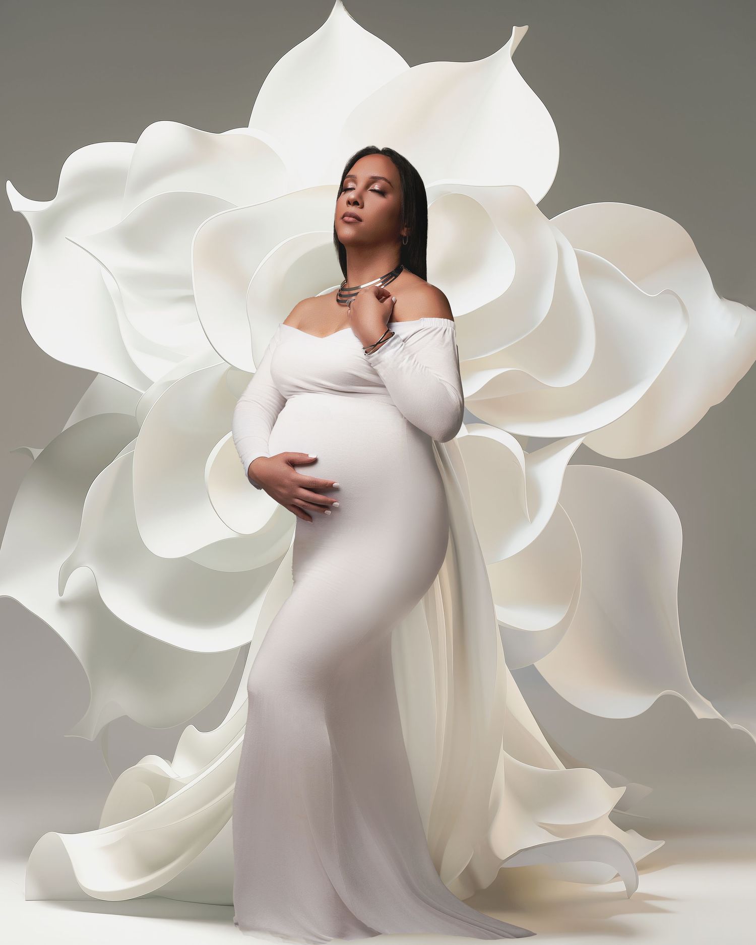 Pregnant woman in white off-shoulder gown poses gracefully against large white floral backdrop in elegant maternity photo.