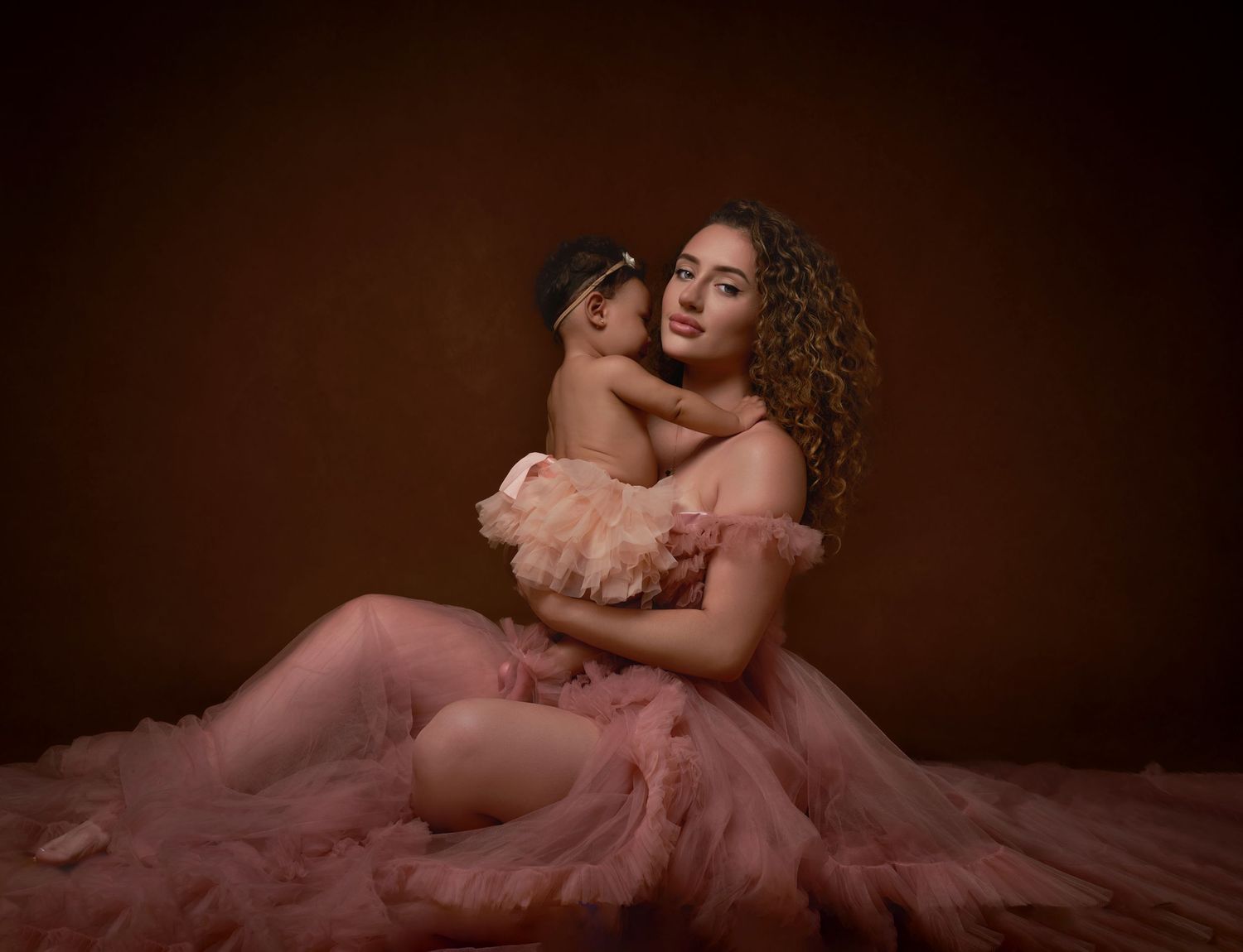 Elegant maternity portrait in soft pink tulle gown with romantic lighting and moody atmosphere.