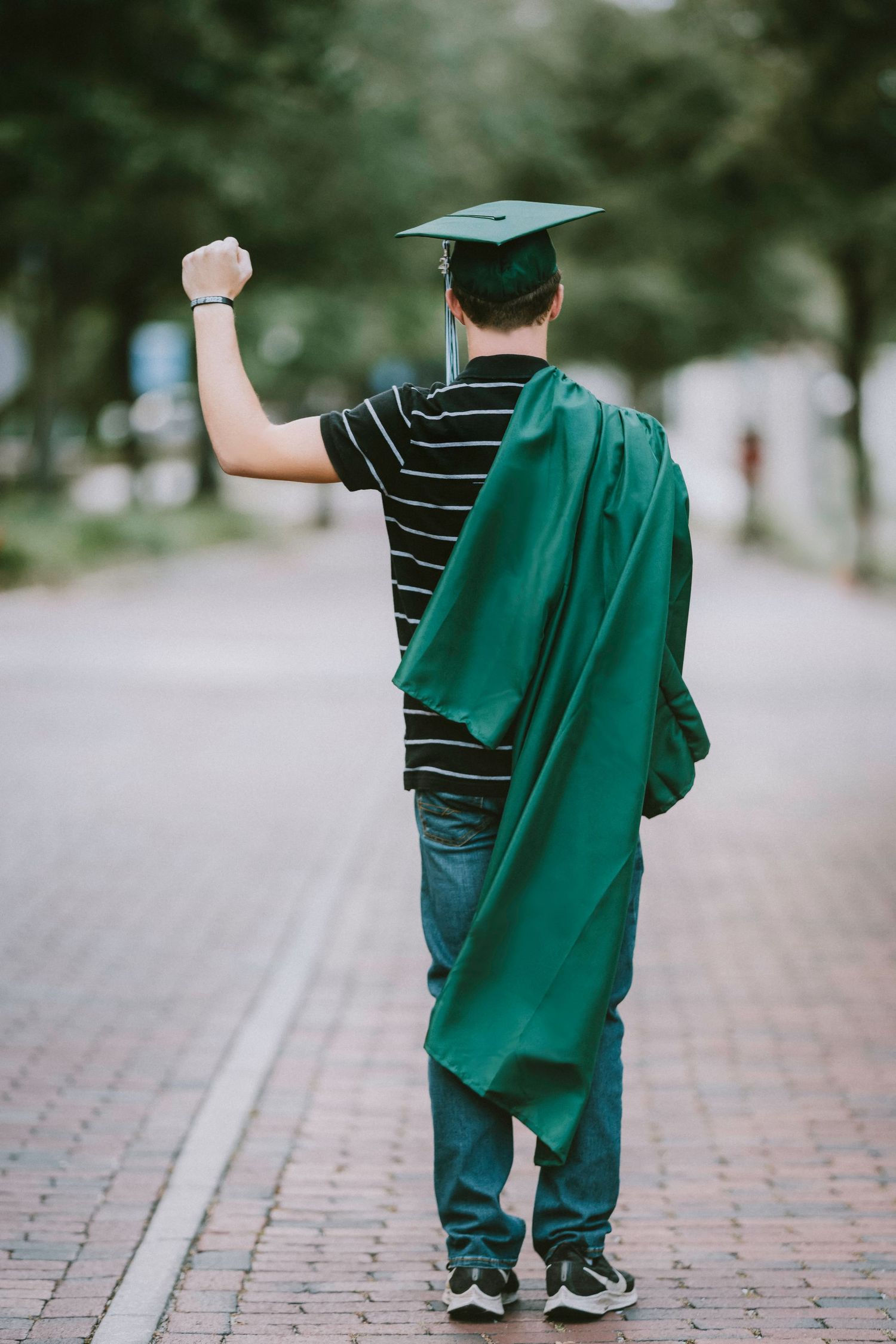 Graduate in green cap and gown walks away on brick pathway walking away.