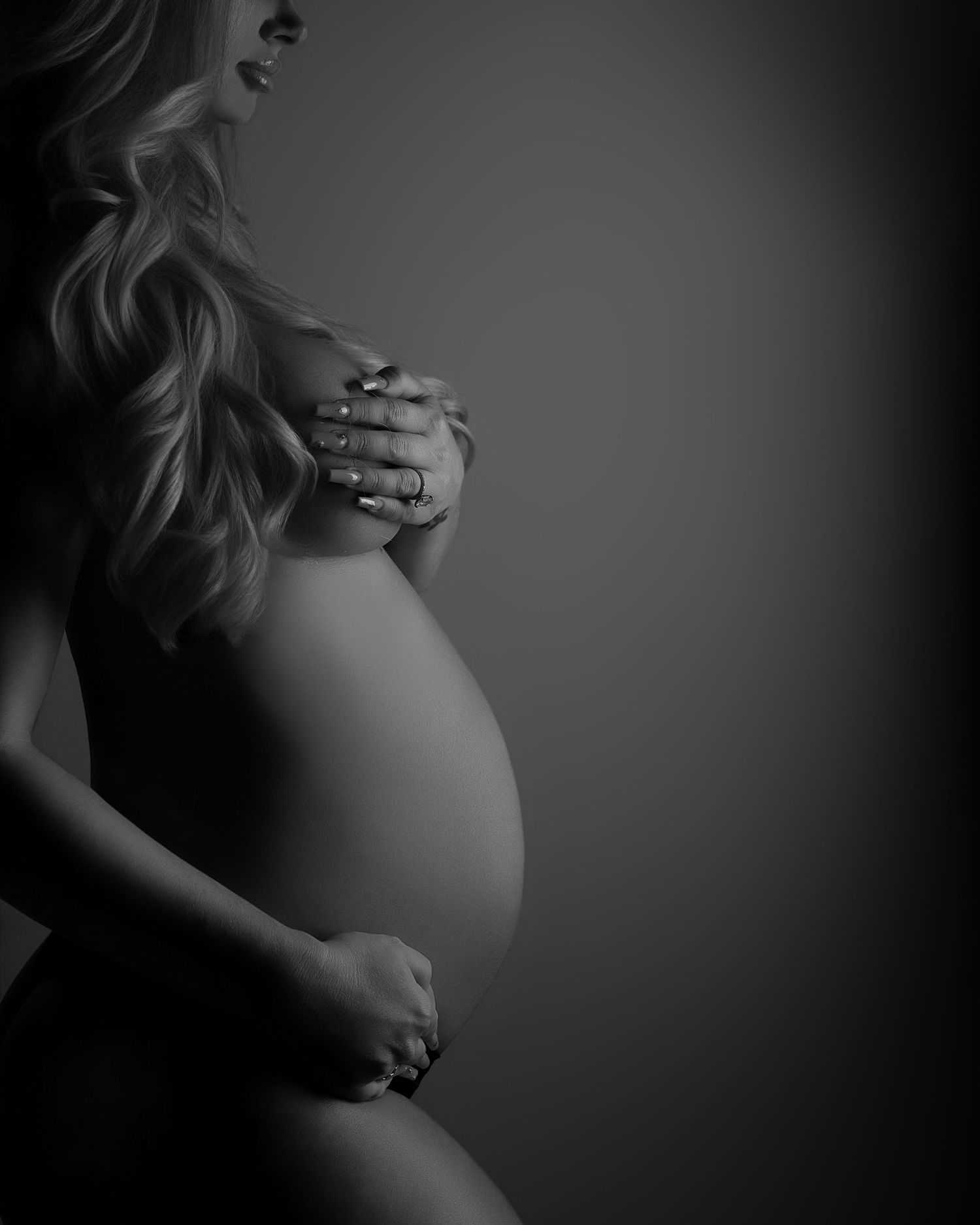 Black and white artistic maternity photography highlighting silhouette and belly against dark background.