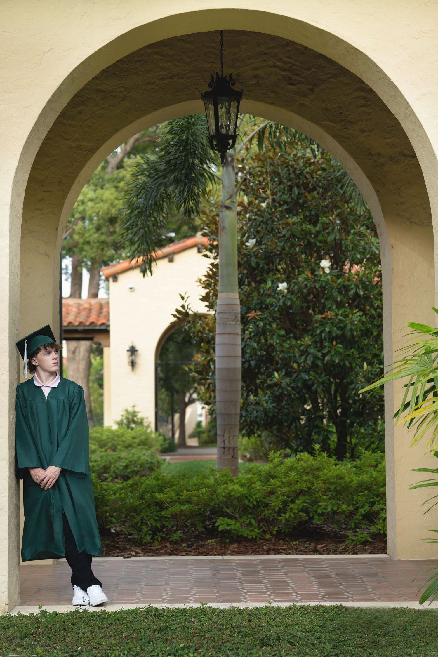 Graduate in green cap and gown poses under arched colonnade of Mediterranean-style building.