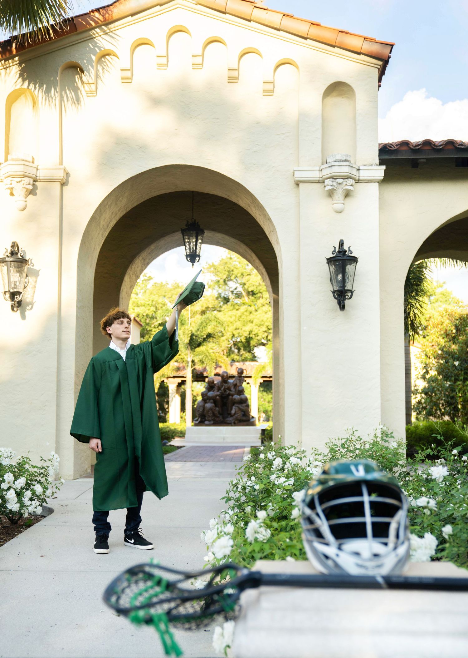 Graduate in green cap and gown poses under arched walkway of Mediterranean-style building.
