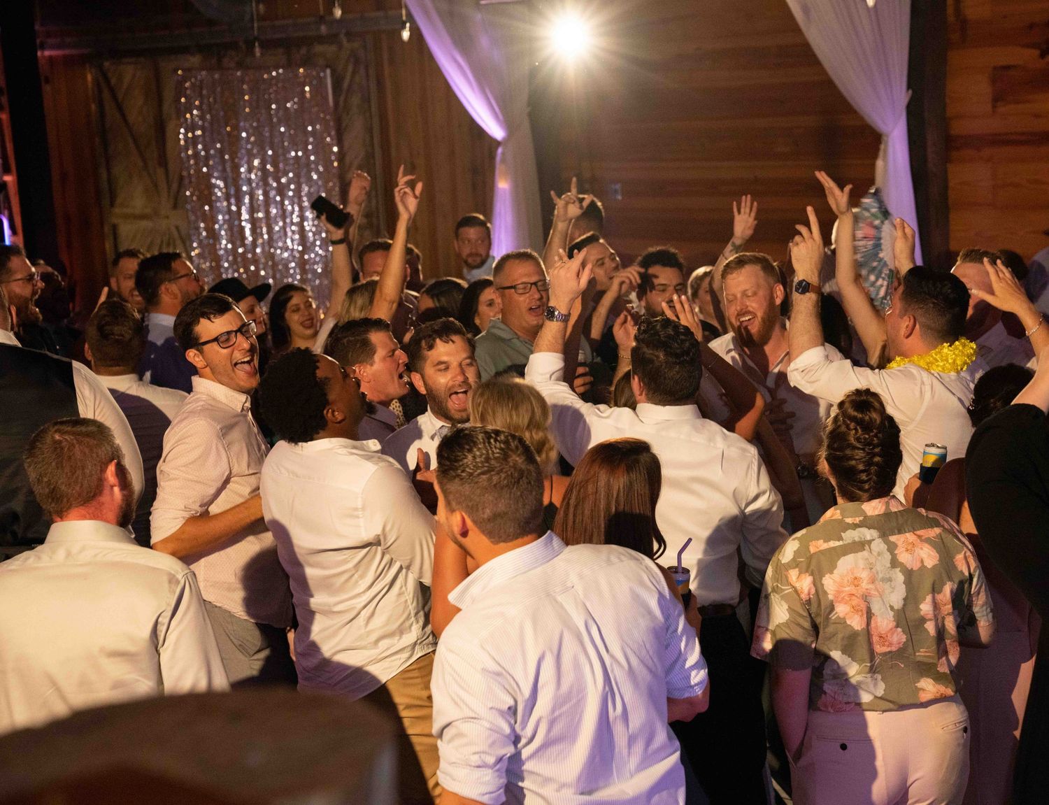 A crowd of wedding guests dancing and celebrating on a dance floor in a rustic barn venue with purple lighting.