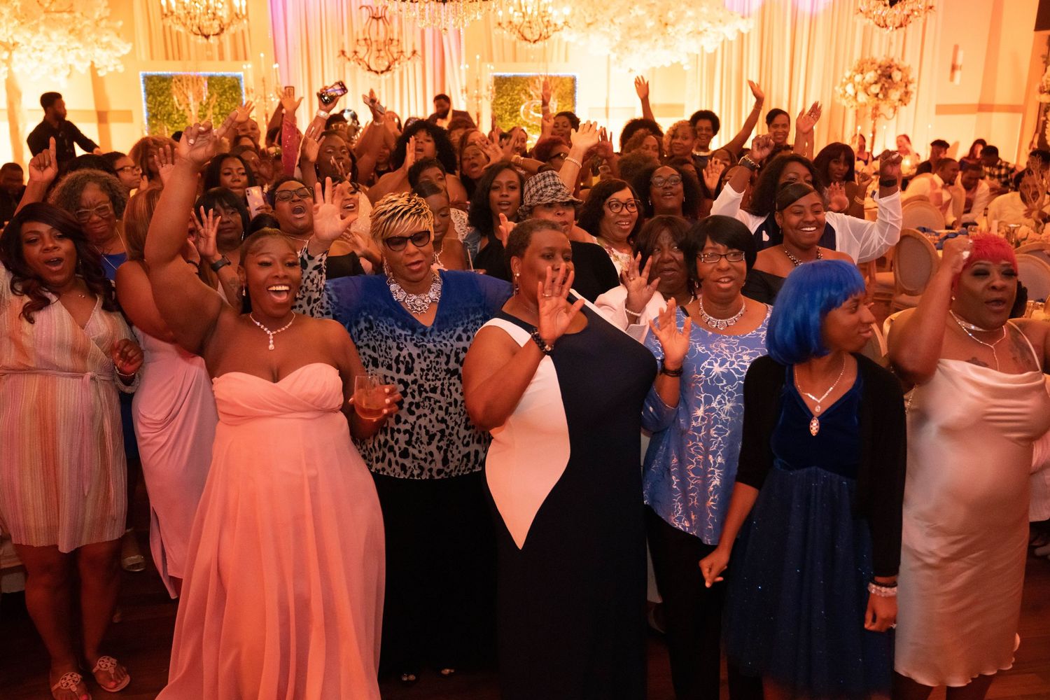 Wedding guests dance and celebrate together on a crowded reception dance floor illuminated by warm lighting.