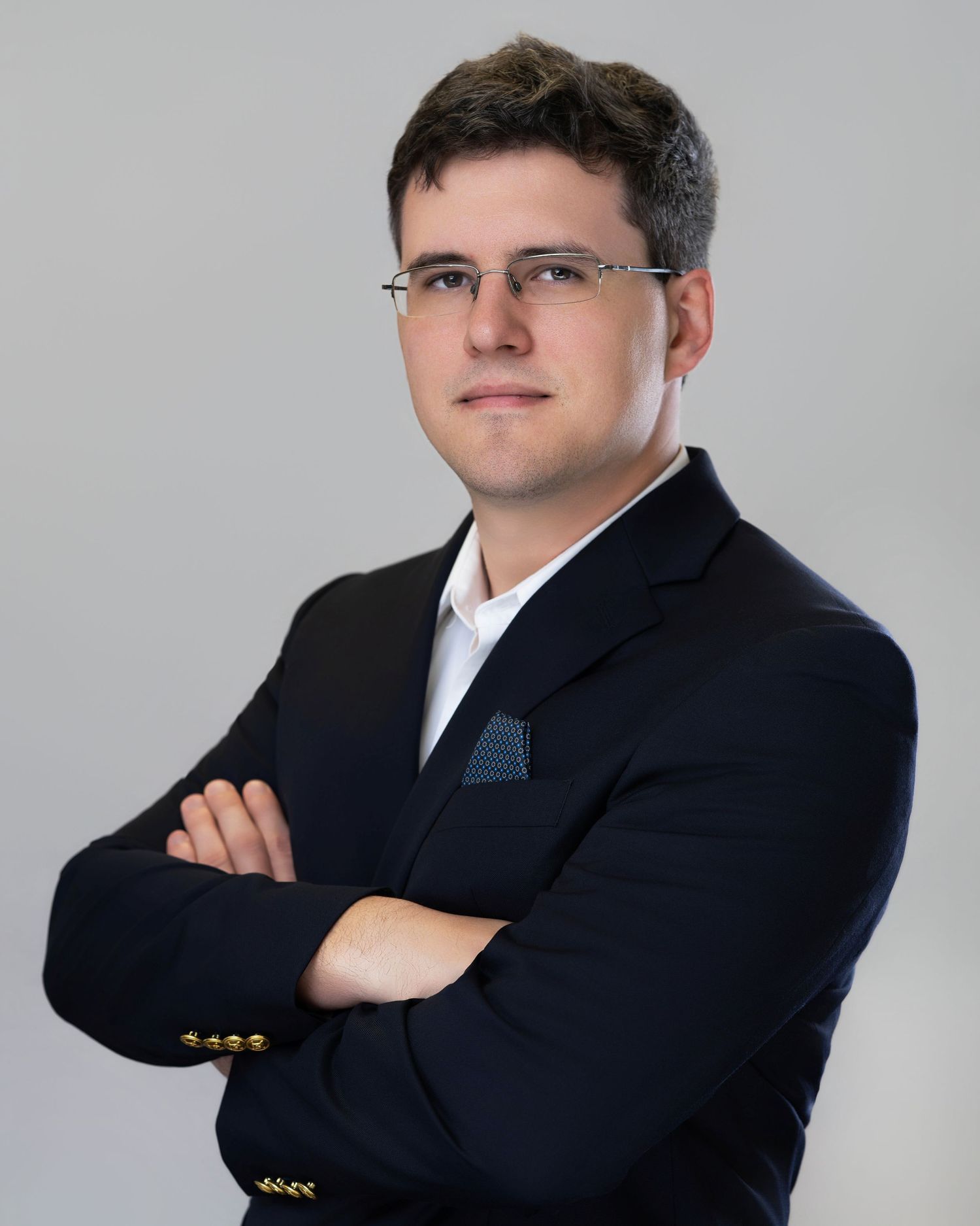 Professional headshot of a person in a dark suit and glasses posing with crossed arms against a gray background.