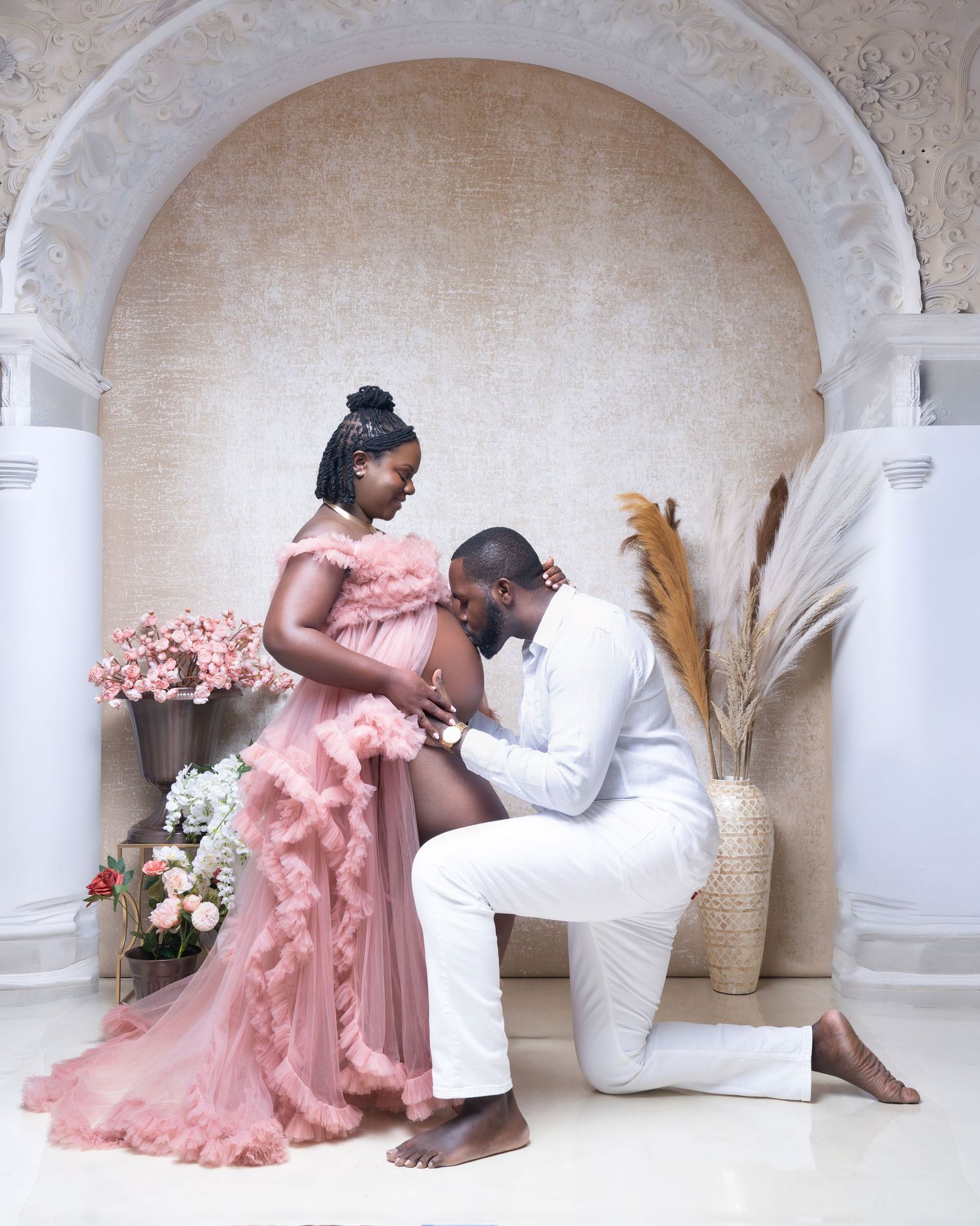 Couple in elegant maternity photoshoot, woman in pink tulle gown, man in white suit kneeling, arched backdrop.
