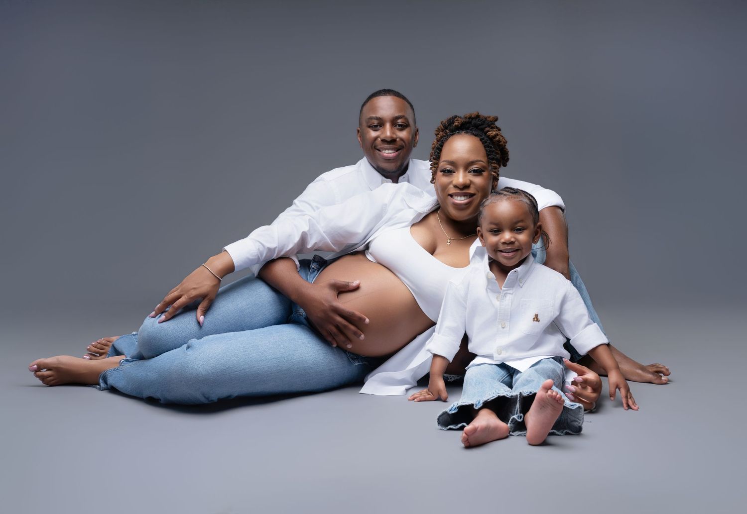 A happy pregnant family posing together in a studio, wearing white shirts and jeans against a gray background.