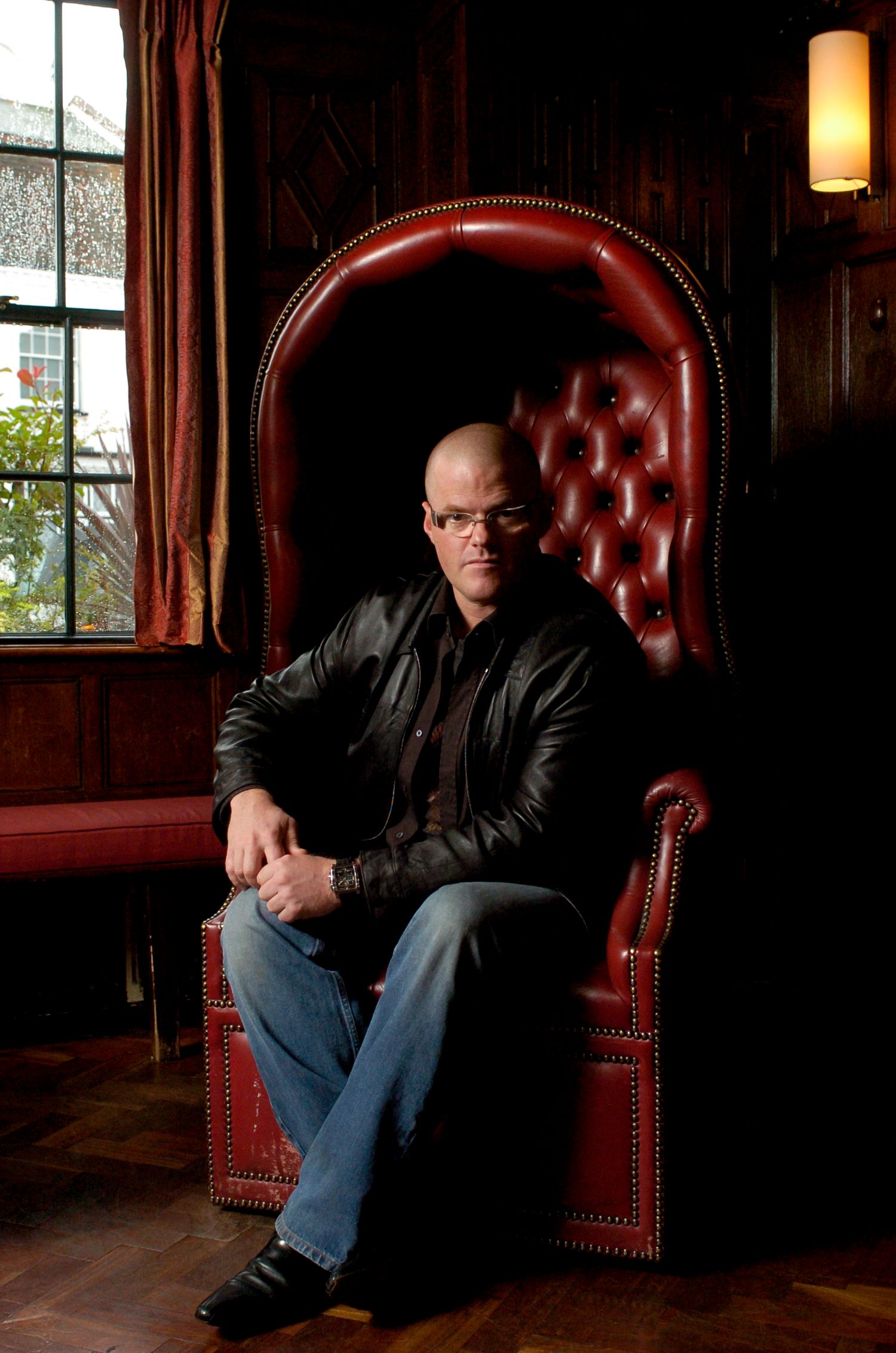 Bald man in black leather jacket sitting in red tufted leather throne chair near window in dark wood interior.