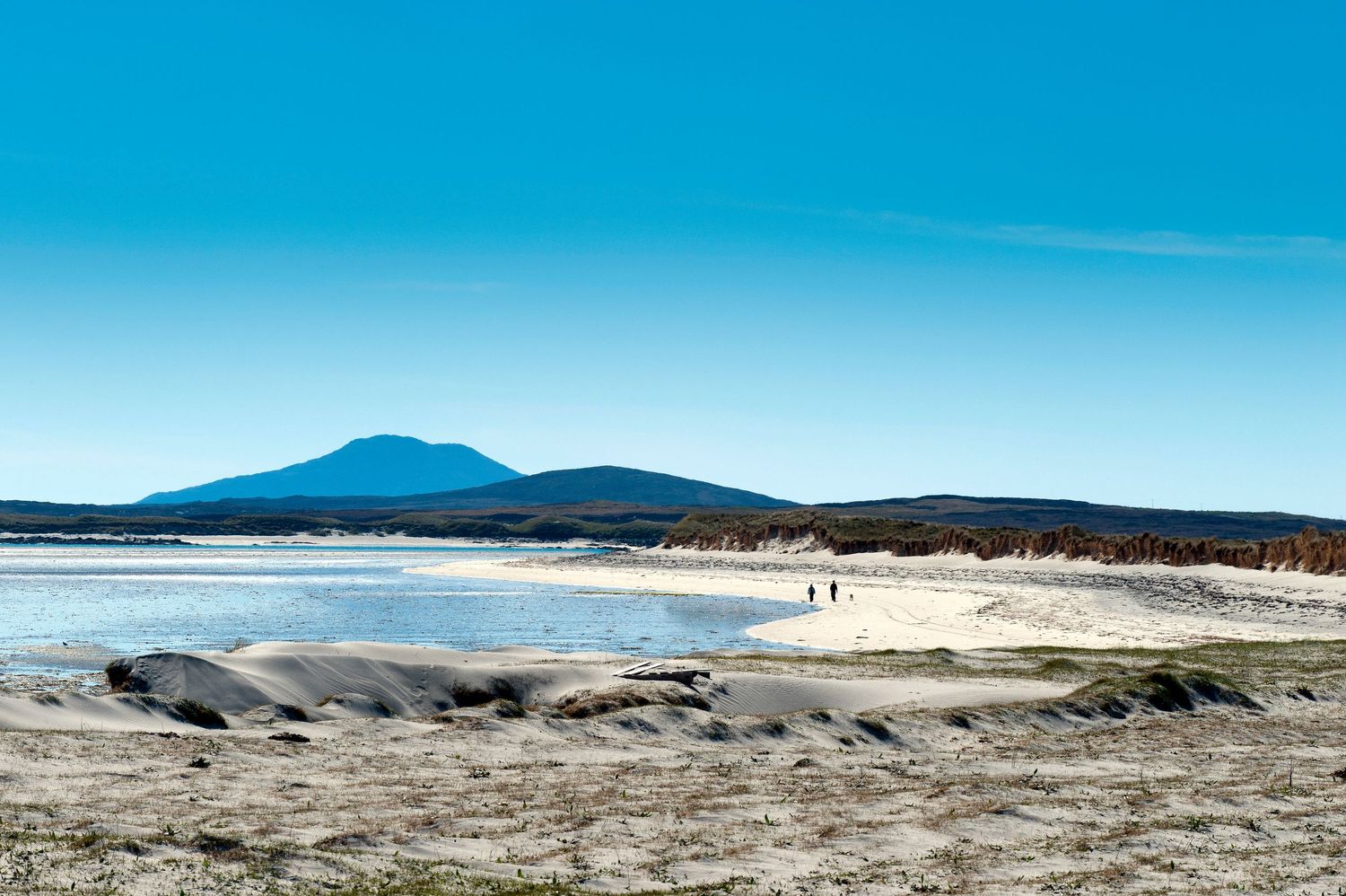 Sandy beach with calm tidal flats, distant mountain peak, and two figures walking under a clear blue sky.