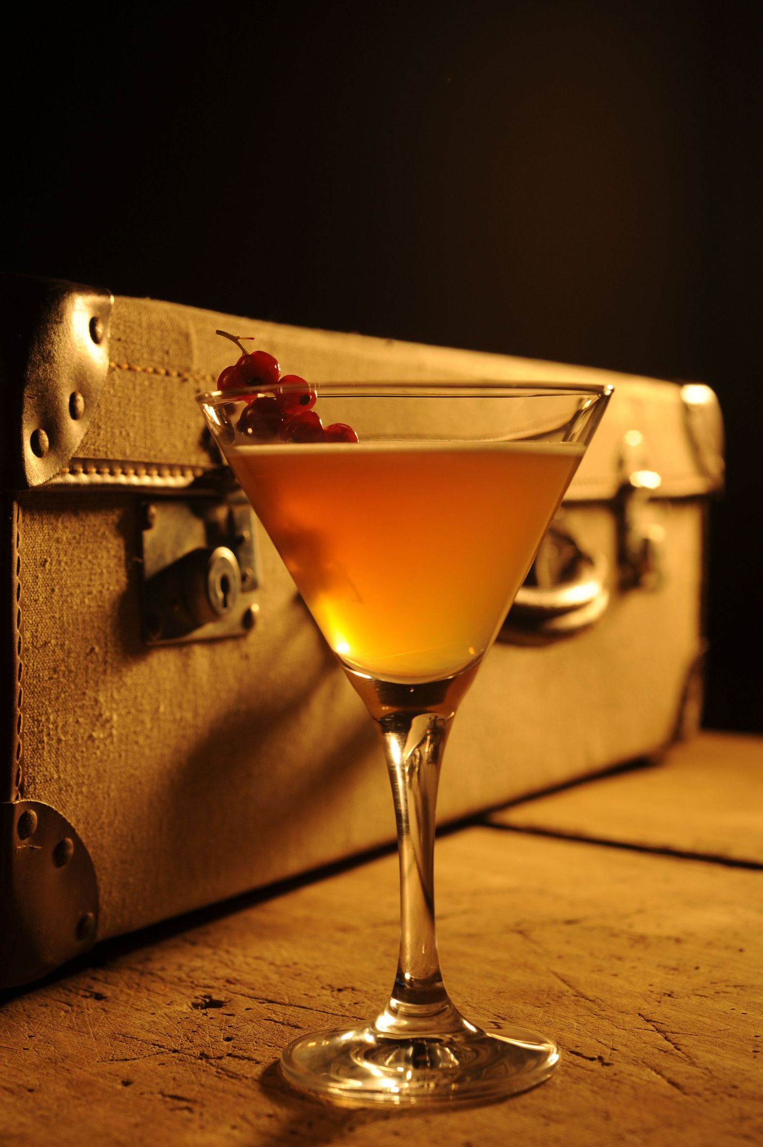 Golden cocktail in martini glass garnished with red berries, set beside a vintage suitcase on a wooden surface.