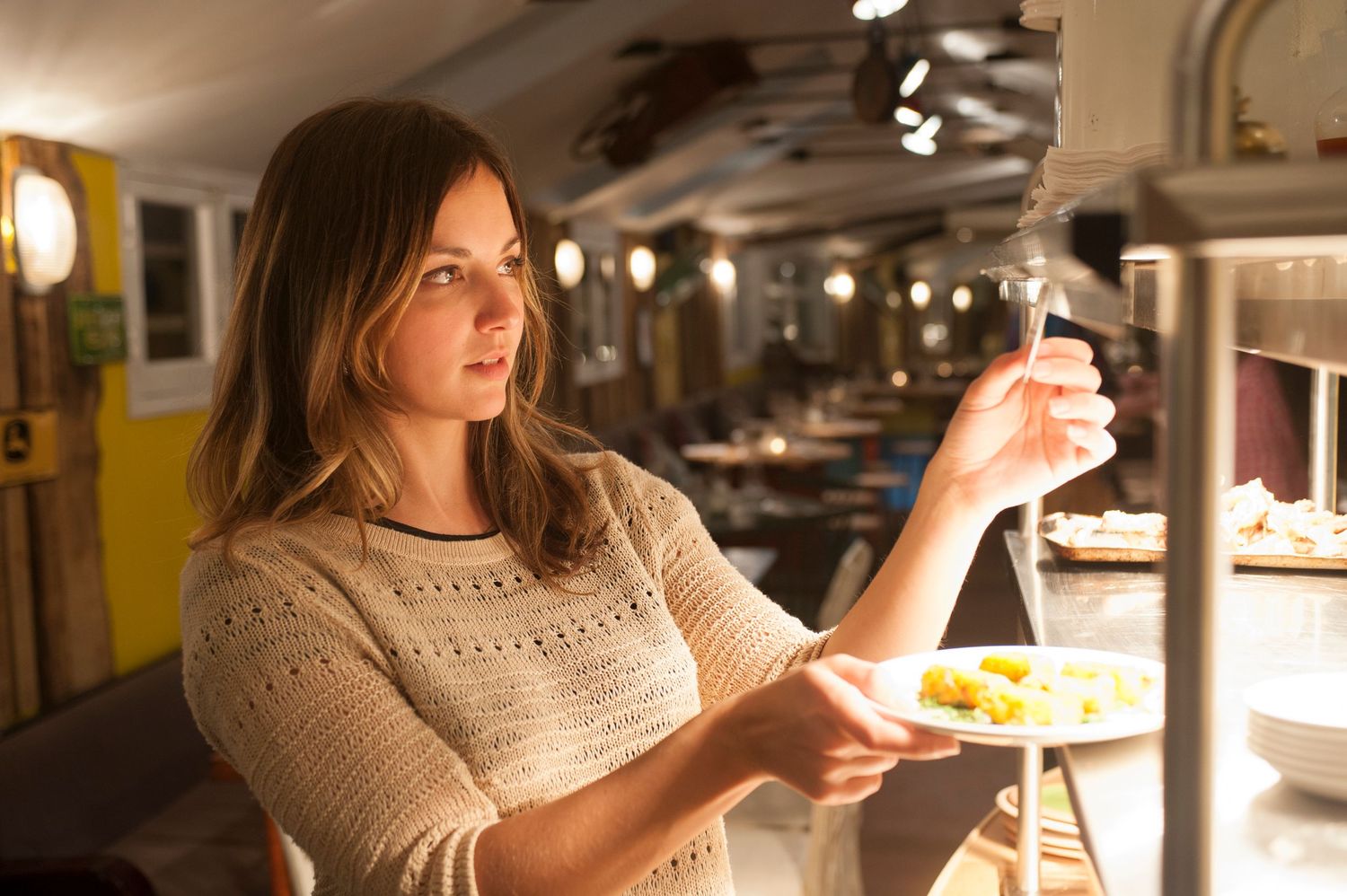 Young woman serving herself food at a buffet restaurant, holding a plate with yellow food under warming lamps.