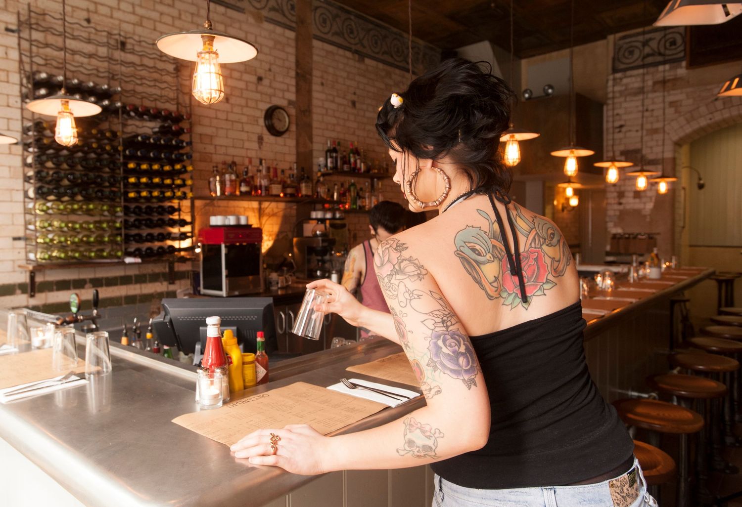 Tattooed female bartender working behind a rustic brick bar with warm Edison bulb lighting and wine storage.