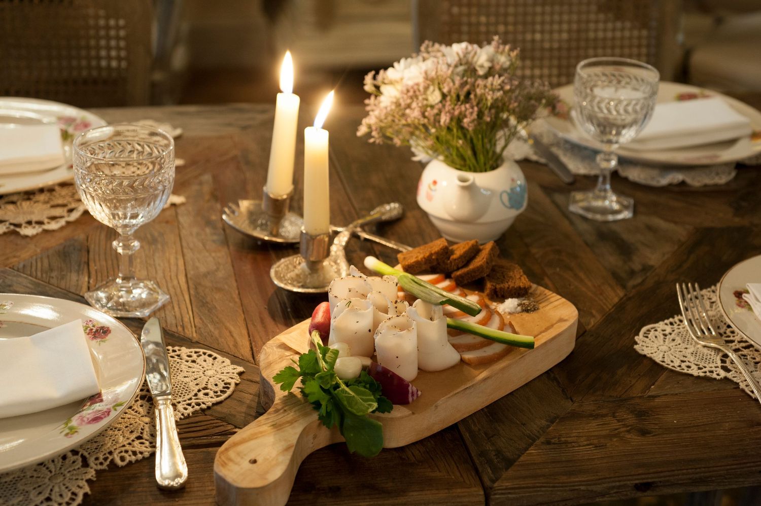 Elegant candlelit dining table with charcuterie board, crystal glasses, white candles, and floral centerpiece on rustic wood.