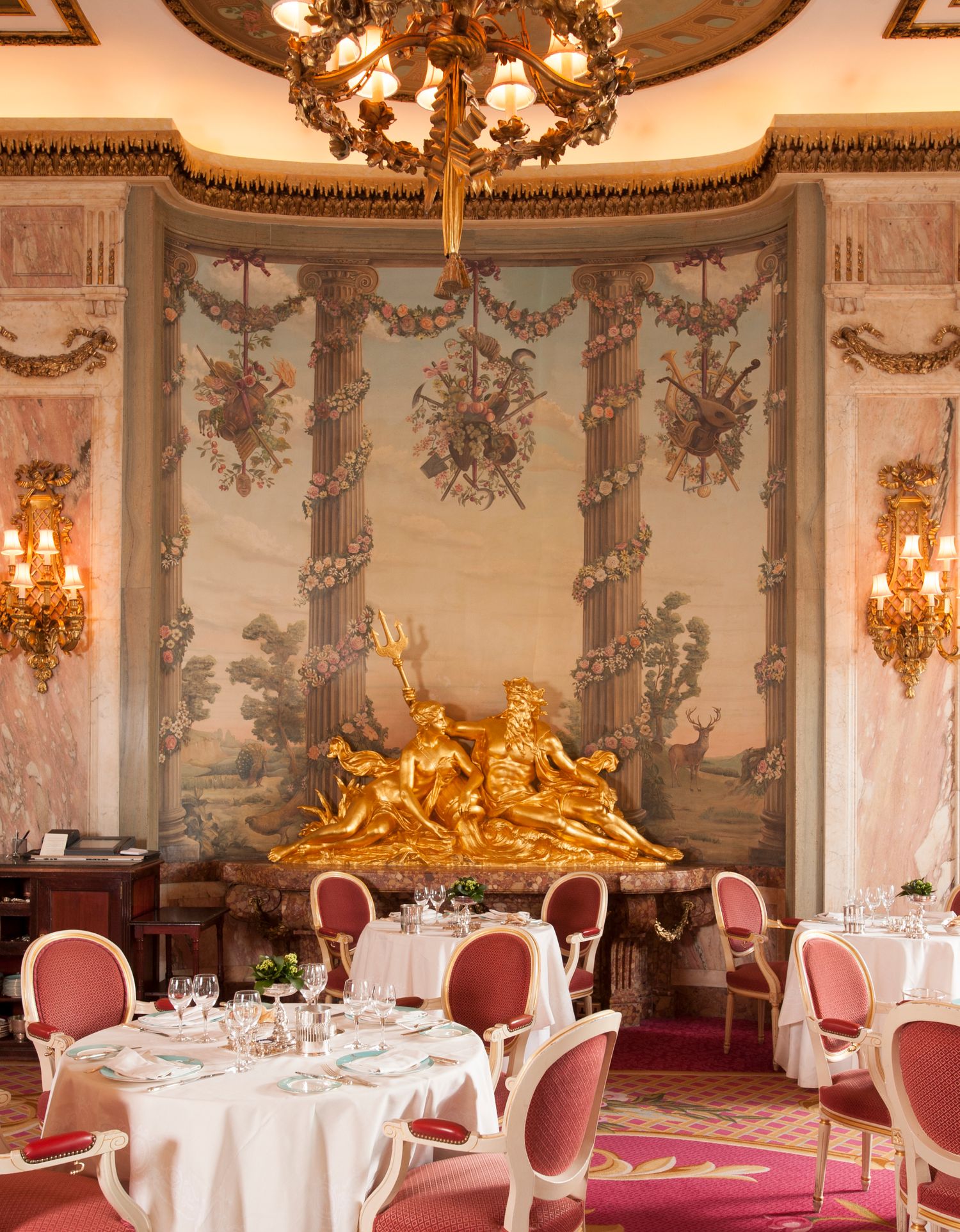 Elegant French dining room with gilded sculpture, ornate tapestry, pink chairs, and chandelier in a luxury restaurant.