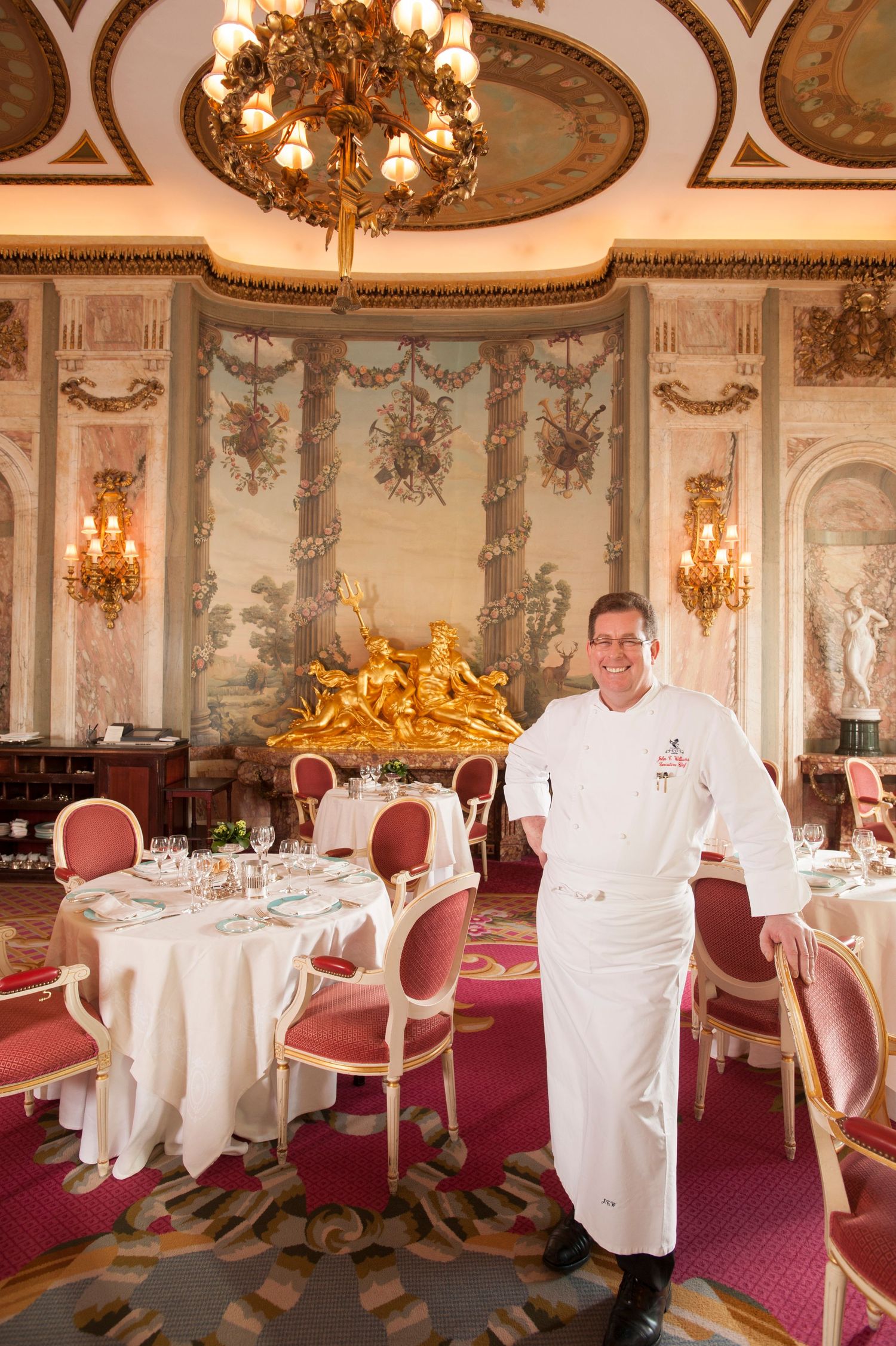 Chef in white uniform poses in elegant dining room with gold chandelier, ornate murals, and formally set tables.