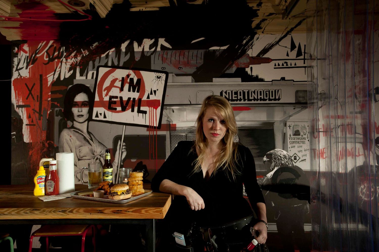 Woman sitting at a table in front of graffiti art mural with 'I'm Evil' text in a dark urban restaurant.