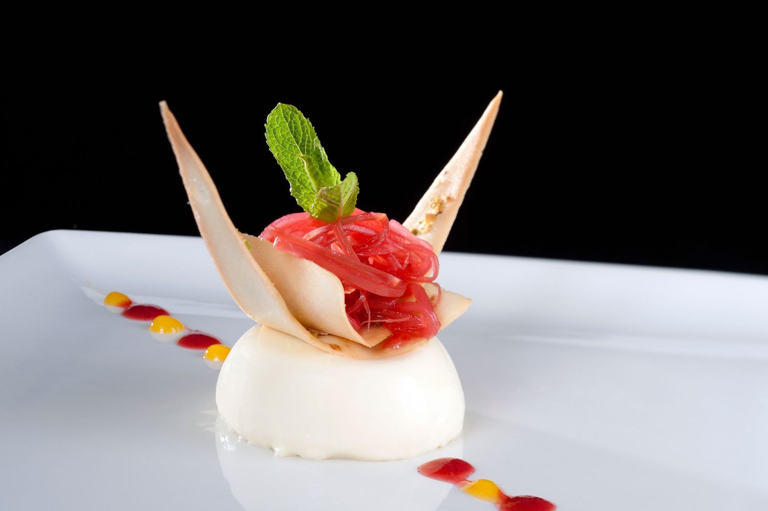 Elegant panna cotta dessert topped with fresh strawberries, crispy tuile, and mint leaf on a white plate.