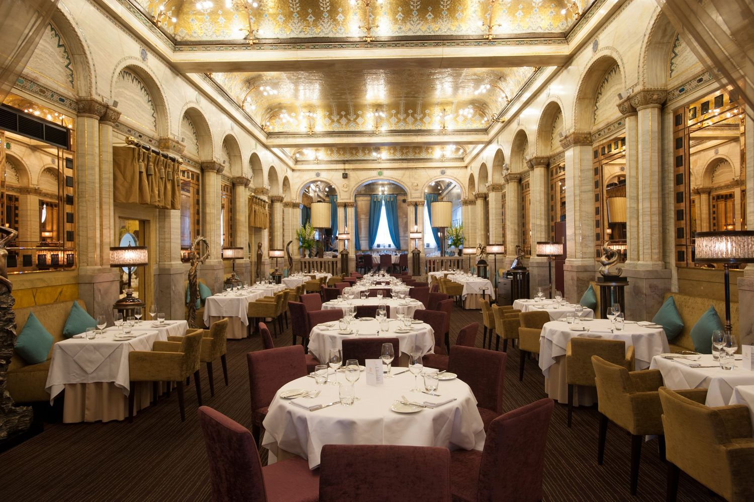 Elegant fine dining restaurant with ornate gold ceiling, white tablecloths, and classical architecture in a grand ballroom setting.