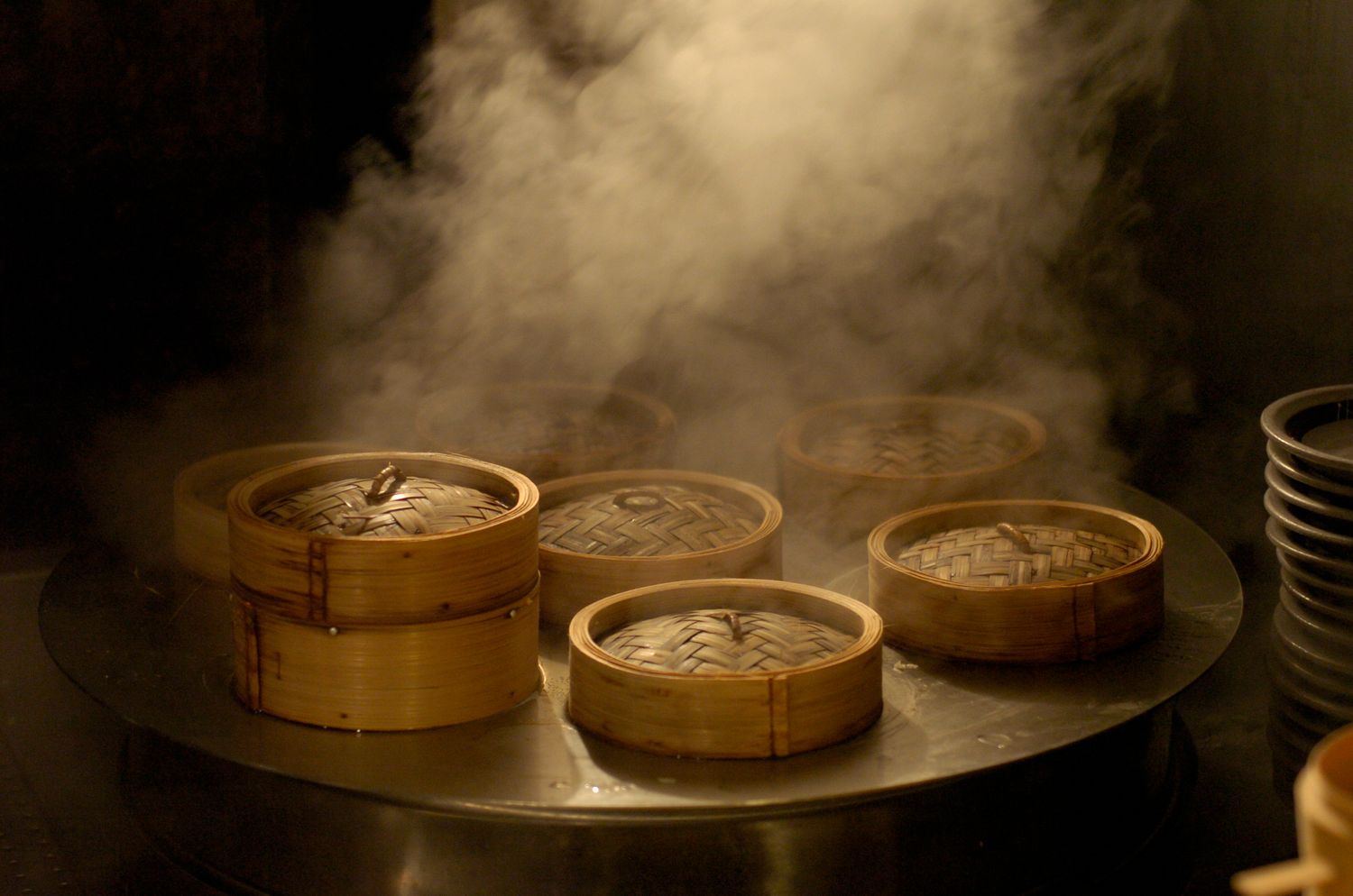 Steaming bamboo dim sum baskets on a metal tray in a dimly lit Chinese restaurant kitchen.