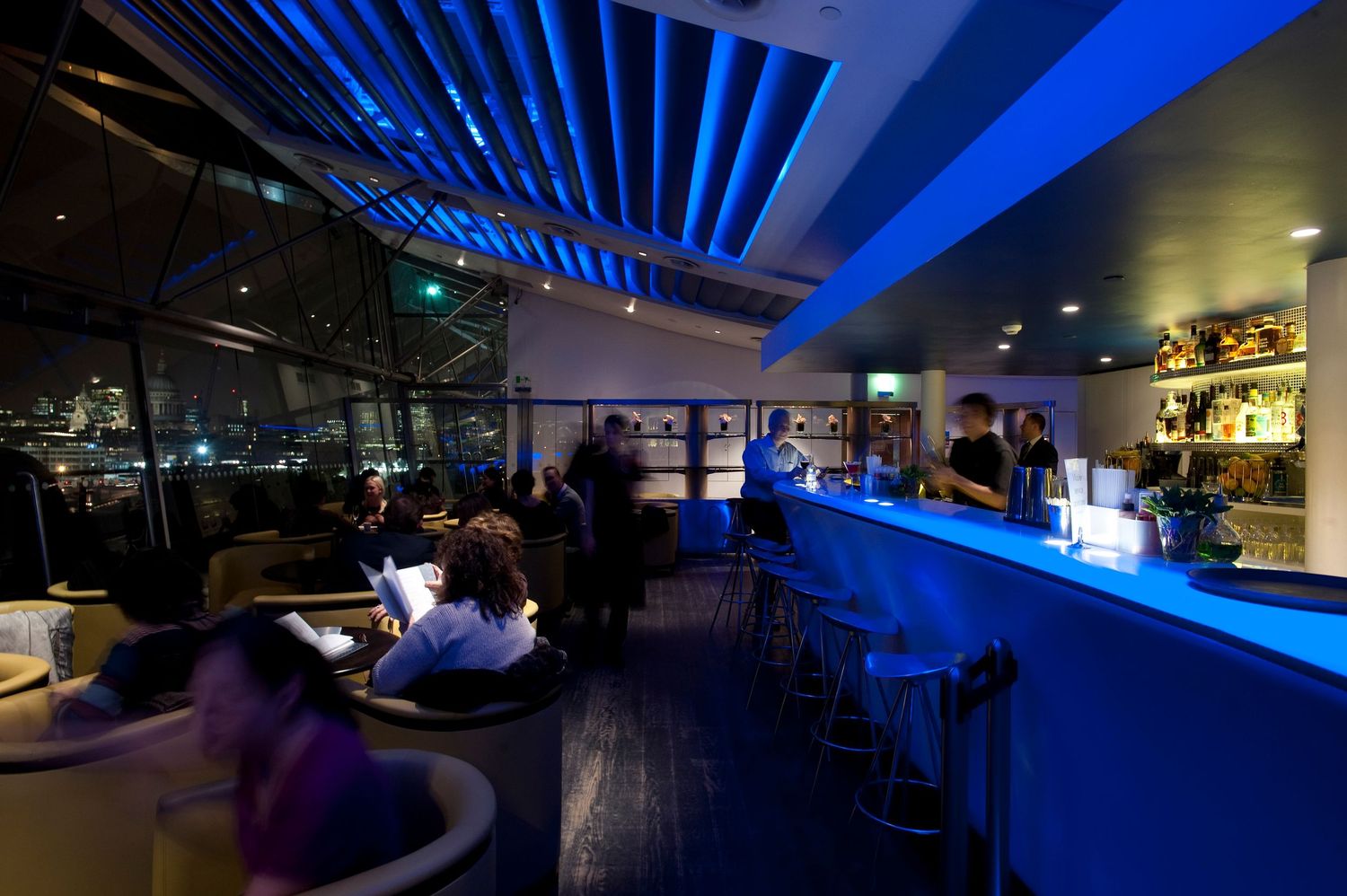 Stylish rooftop bar with blue neon lighting, city views, bar stools, and patrons enjoying evening drinks.