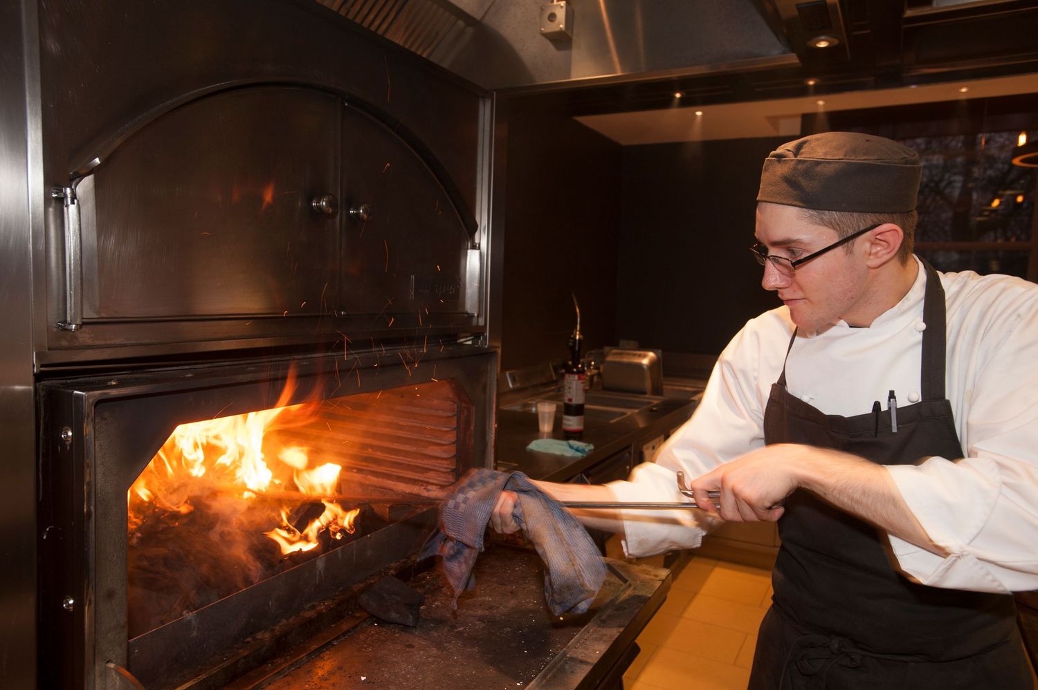Chef tending to wood-fired grill in restaurant kitchen, adding logs to open flame cooker.