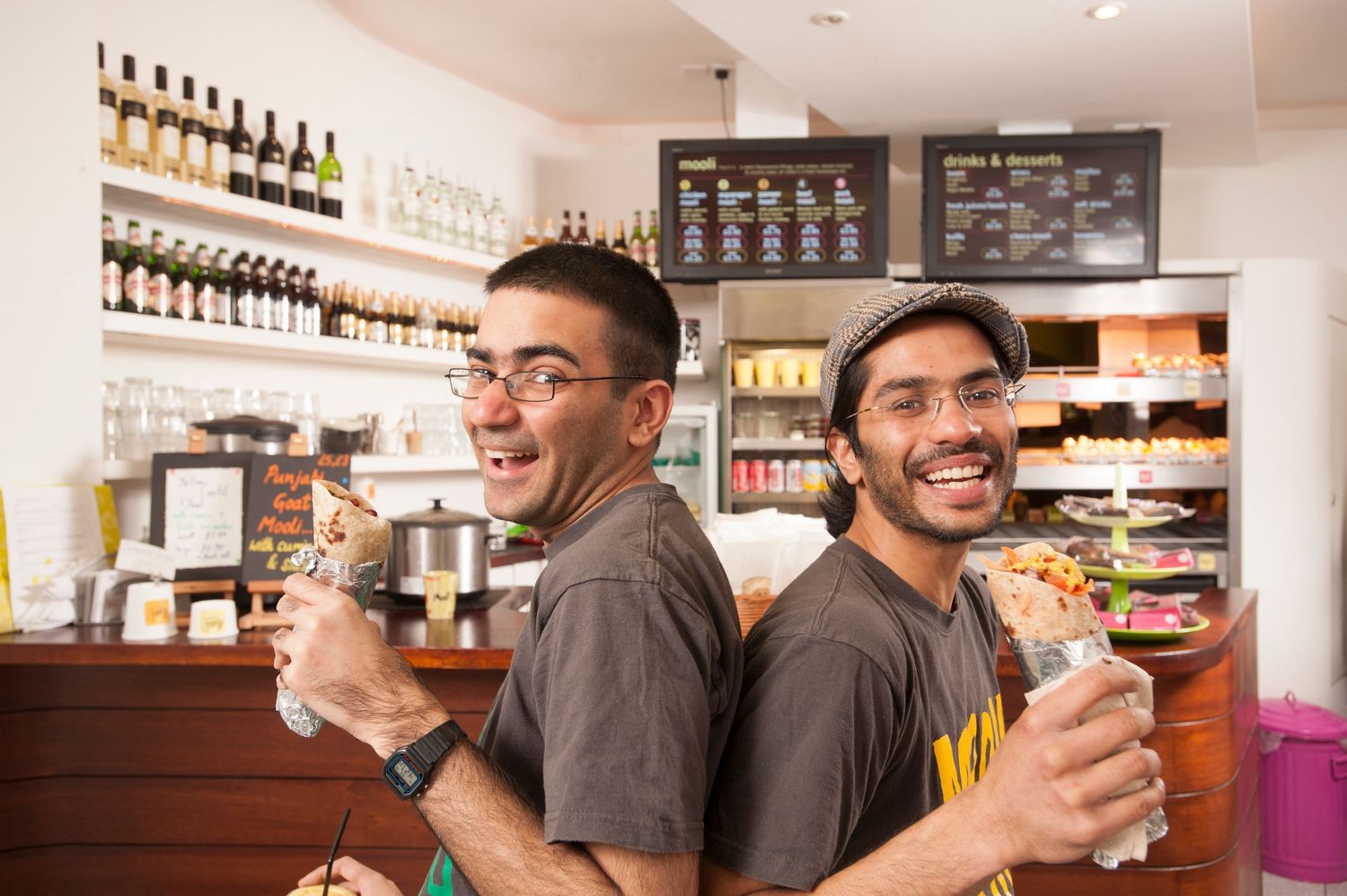 Two smiling men pose back-to-back in a cafe, holding food and drinks near a bar with bottles and menu boards.