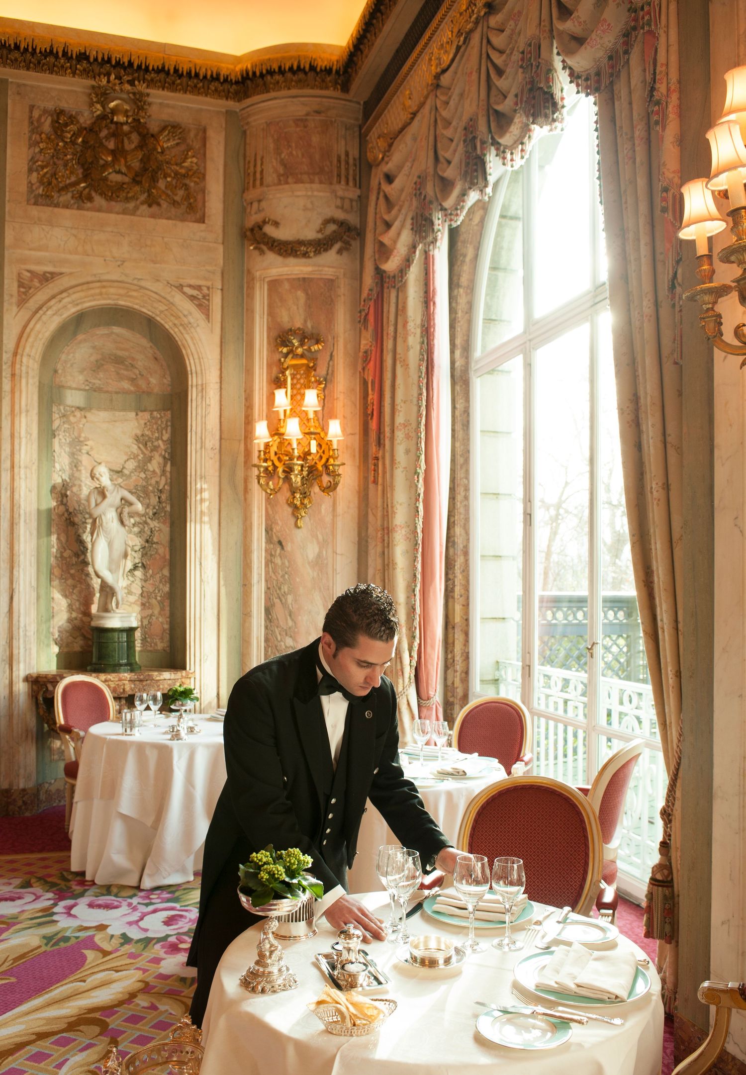 Elegant waiter in tuxedo setting a formal dining table in a luxurious French restaurant with ornate decor.