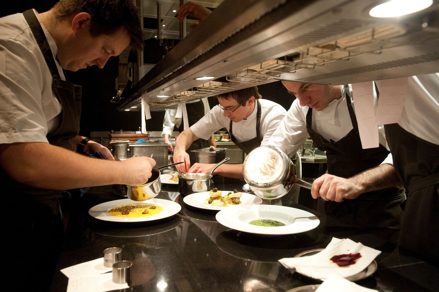 Three chefs in black uniforms carefully plate gourmet dishes in a professional restaurant kitchen.