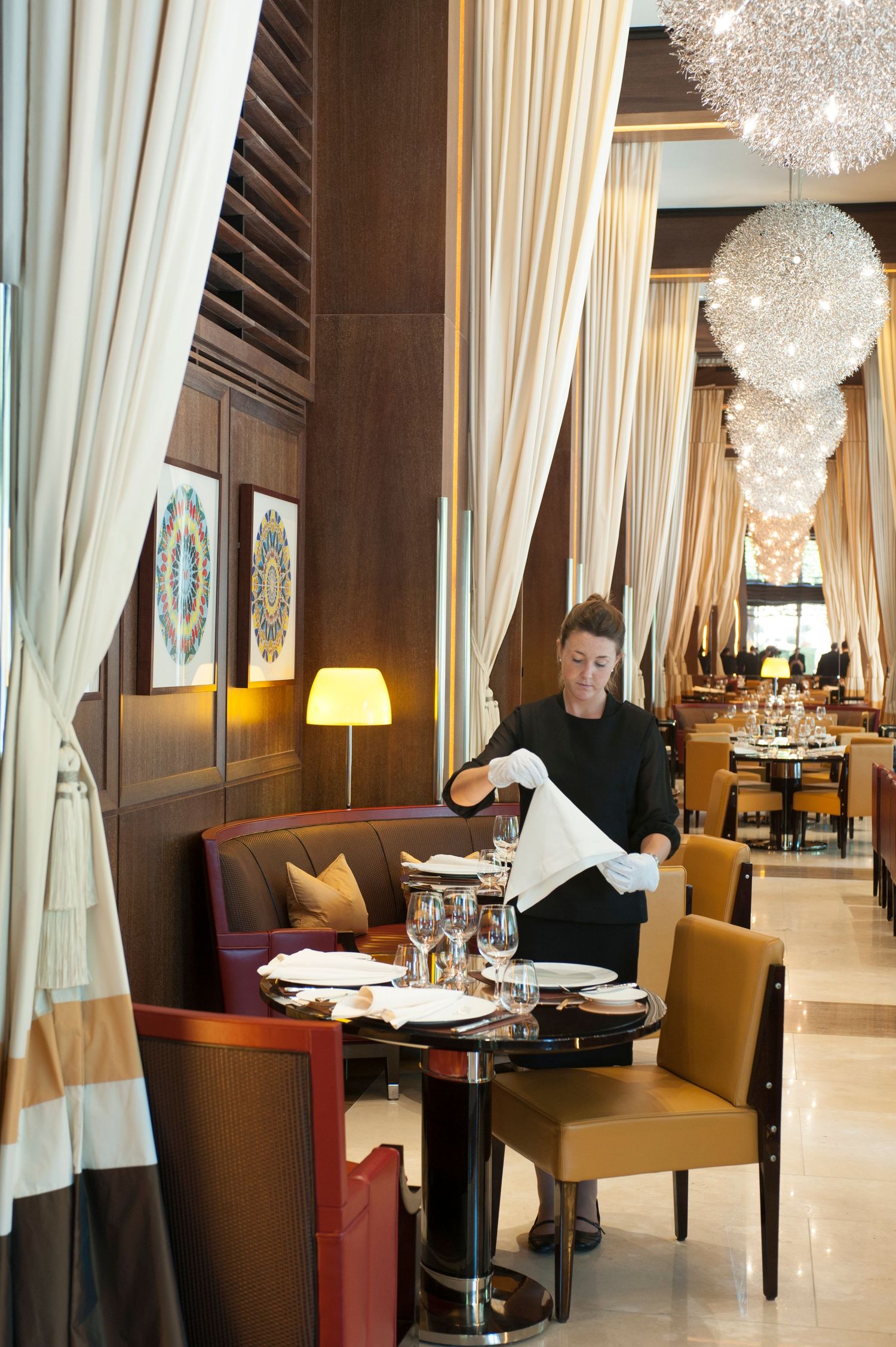 Elegant restaurant interior with a server setting tables near decorative pendant lights and warm wood paneling.