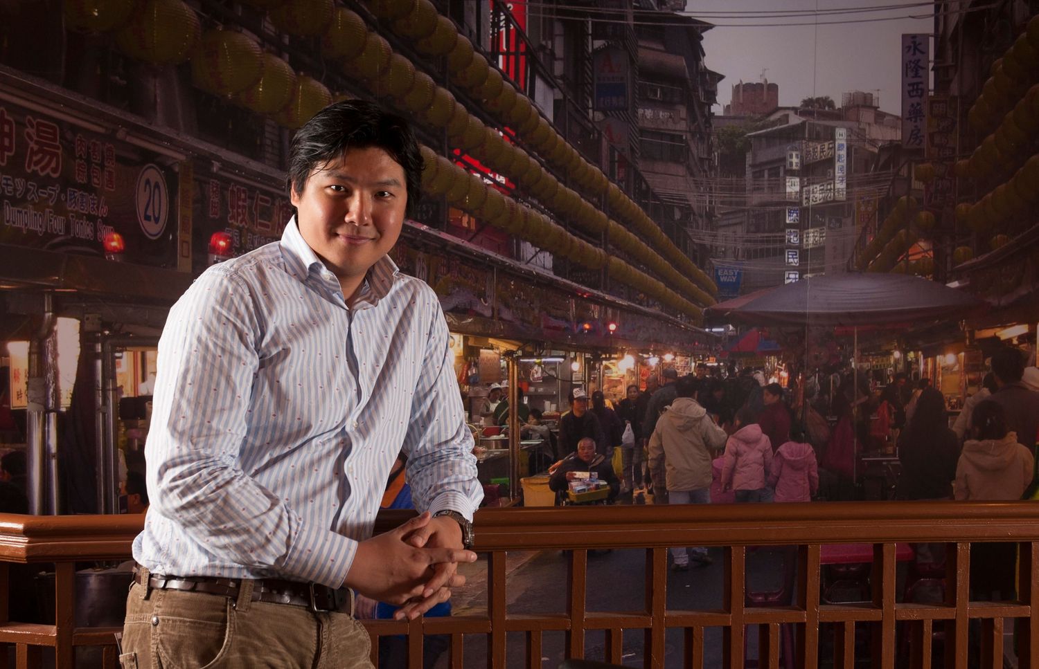 A man poses confidently in front of a vibrant Asian night market with colorful lights and bustling crowd.