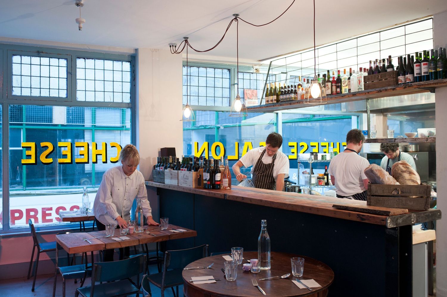 Interior of a cheese salon restaurant bar with staff working behind a wooden counter, wine bottles on shelves, and large windows.