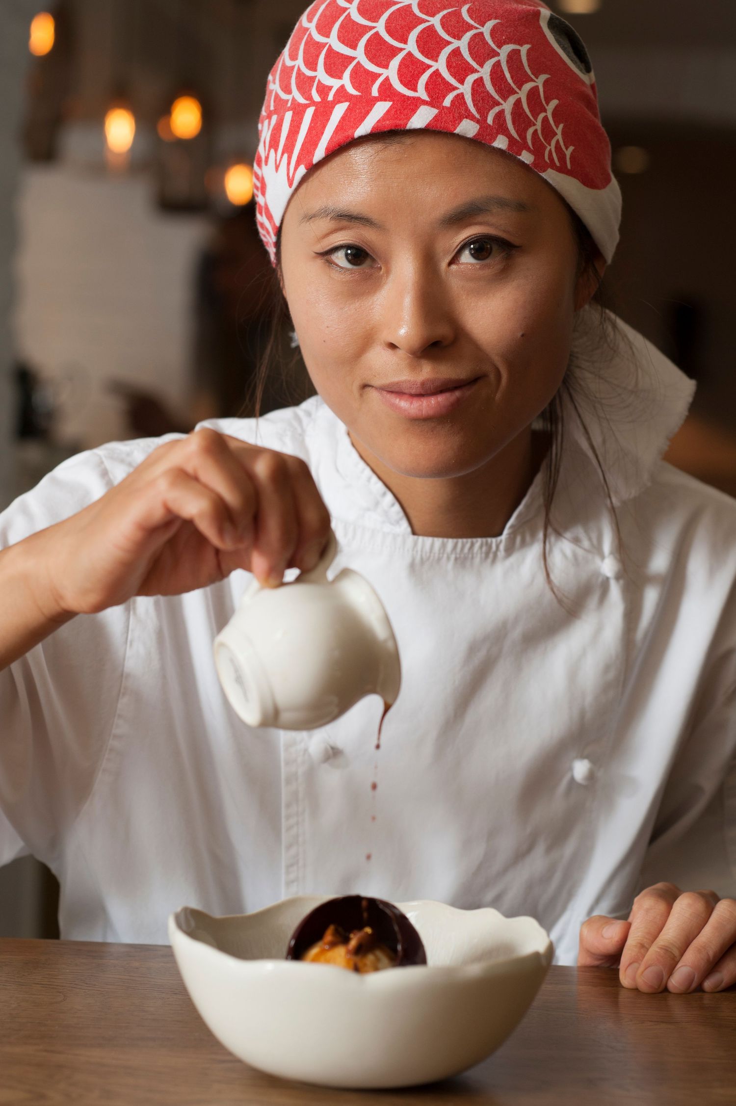 Female chef in white coat and red bandana pouring sauce over a dessert in a white bowl.